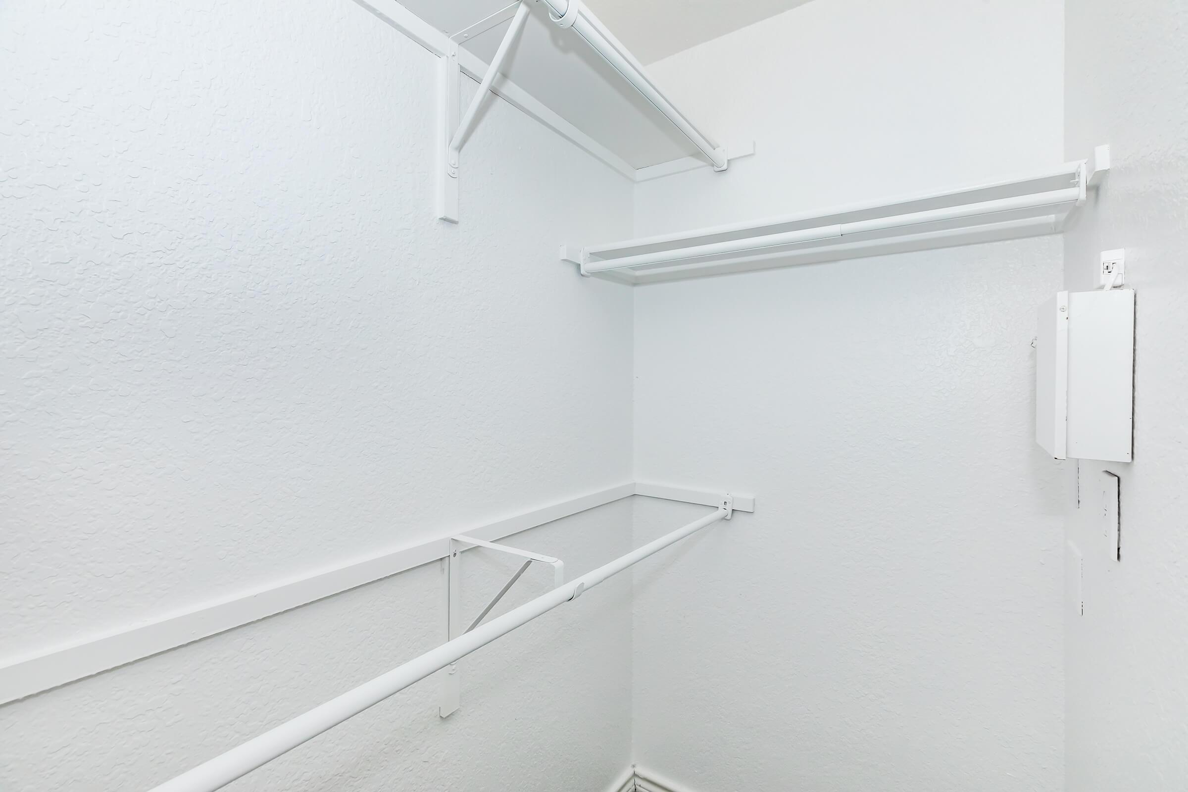 Empty closet with white walls and shelving. The space features two horizontal bars and additional shelving, all in white, creating a clean and minimalistic appearance. There is a small white electrical panel on one side.