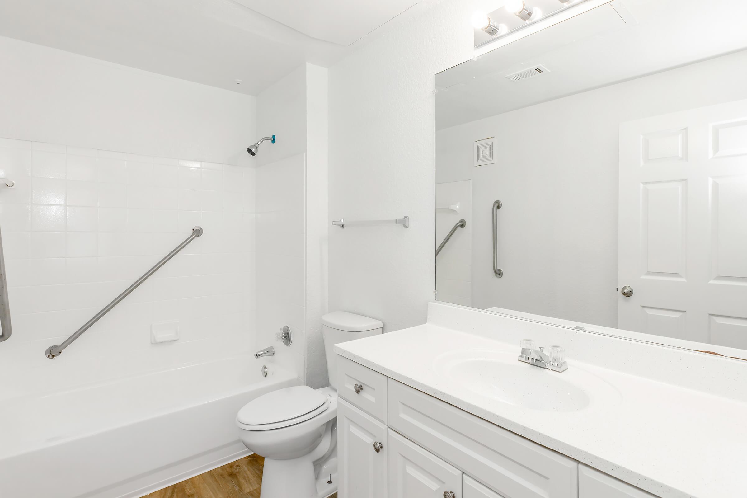 A clean, minimalistic bathroom featuring a white bathtub with shower, a single sink vanity with a countertop, a toilet, and a large mirror. The walls are painted white, and there's a towel bar and grab bars installed for safety. The floor has a light wood finish.