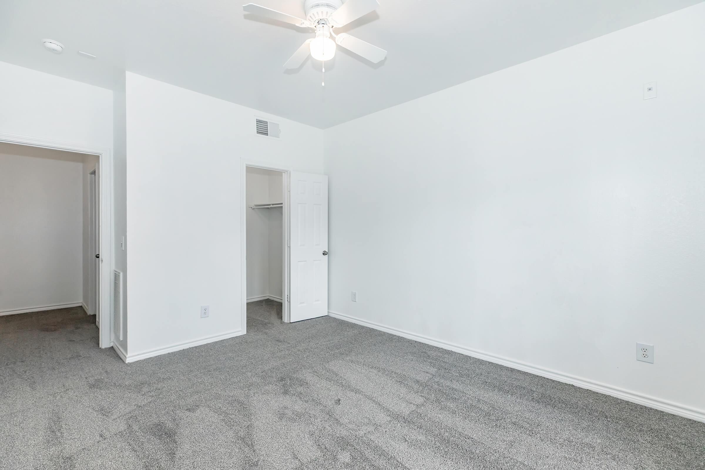 Empty room with light gray carpet, white walls, and a ceiling fan. There are two doorways: one leads to a closet and the other to a hallway. Natural light brightens the space, creating a clean and airy atmosphere.