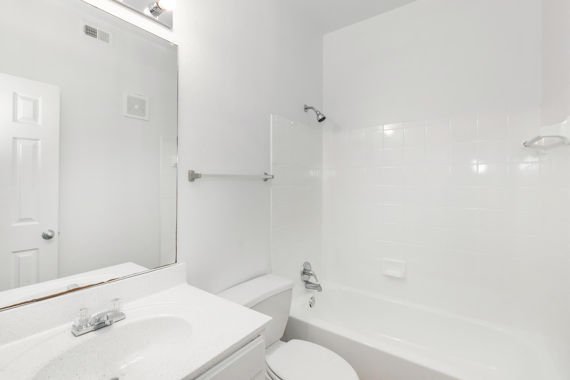A clean, modern bathroom featuring a white bathtub, toilet, and sink with a countertop. There is a large mirror above the sink and white tiled walls, creating a bright and spacious feel. A showerhead is mounted on the wall above the bathtub, and a towel bar is visible. The door is closed, indicating privacy.