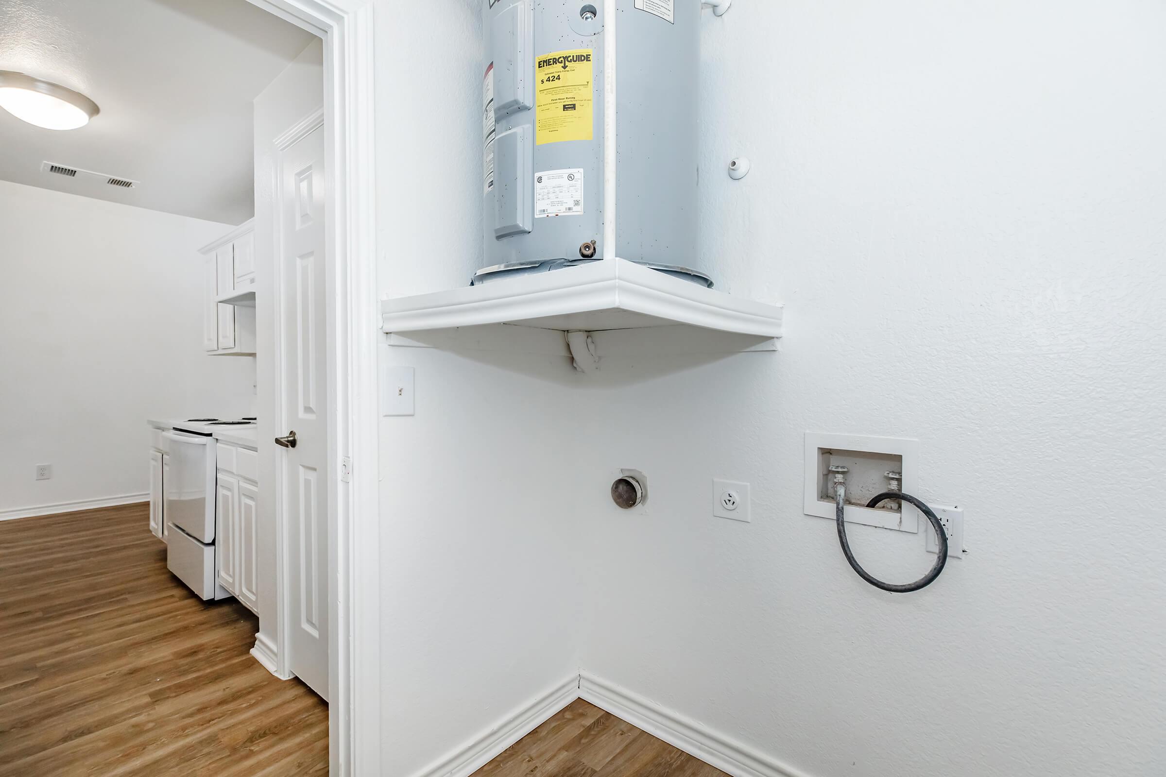 A small laundry area featuring a water heater mounted on the wall, with a washing machine hookup socket nearby. The room has light-colored walls and a wooden floor, with a glimpse of a kitchen area in the background. The space is well-lit and organized.