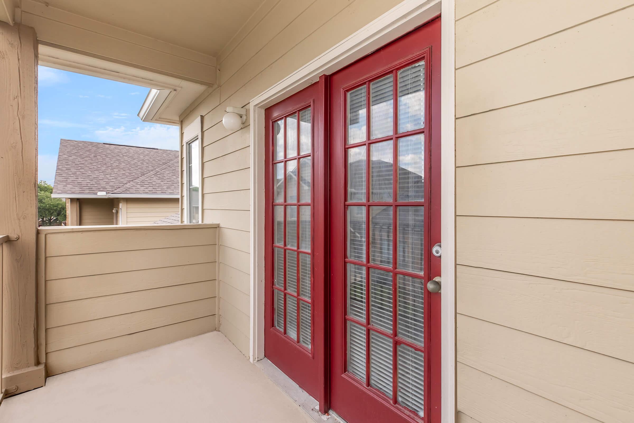A spacious balcony featuring double red French doors with multiple glass panels, leading to an exterior with light-colored siding. The area is well-lit with natural light and offers a view of nearby rooftops and blue sky.