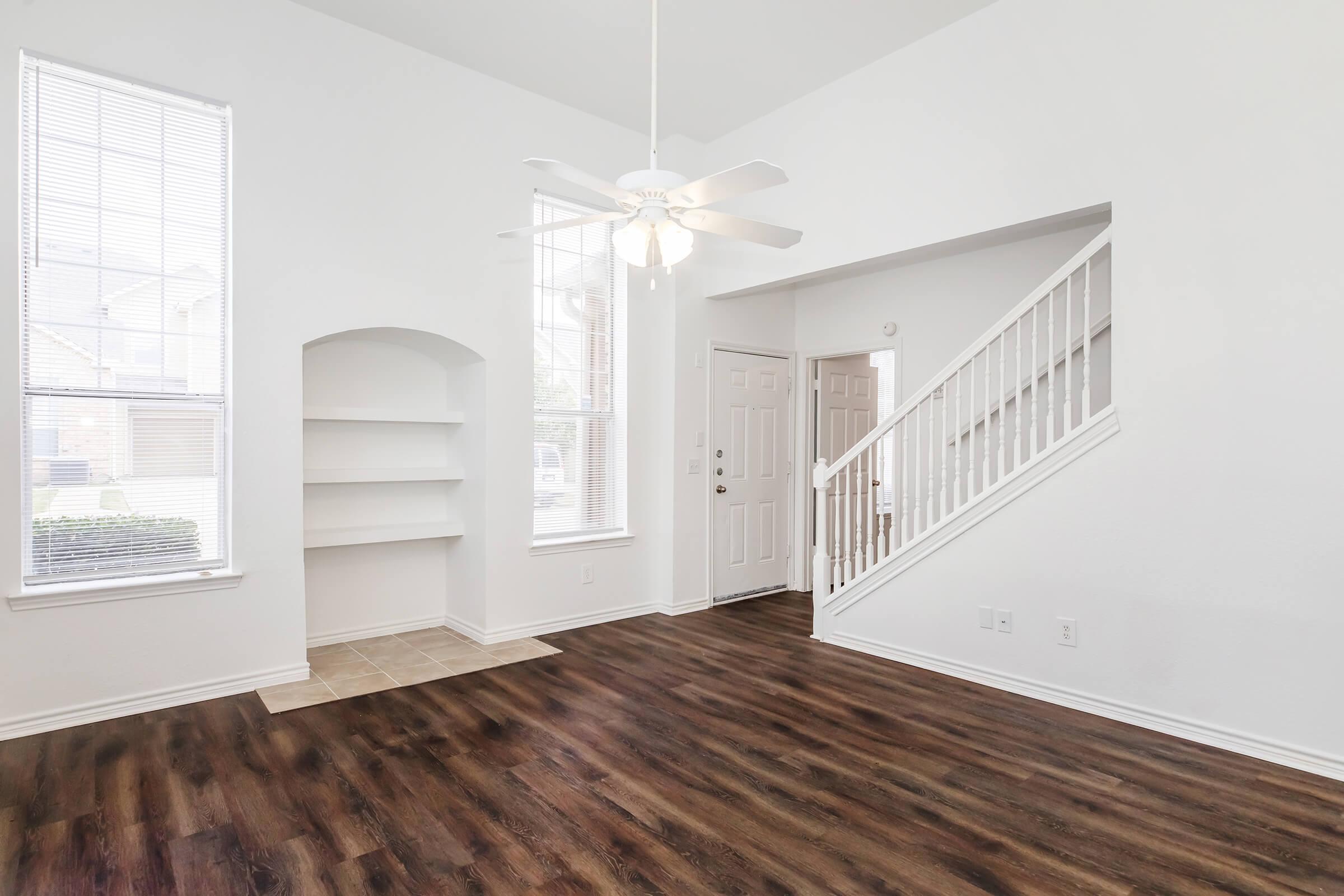 A spacious living area with high ceilings, featuring large windows that let in natural light. The room includes a ceiling fan, a built-in shelving unit, and an entrance with a staircase leading to the upper level. The flooring is dark wood, and the walls are painted white, creating a bright and airy atmosphere.