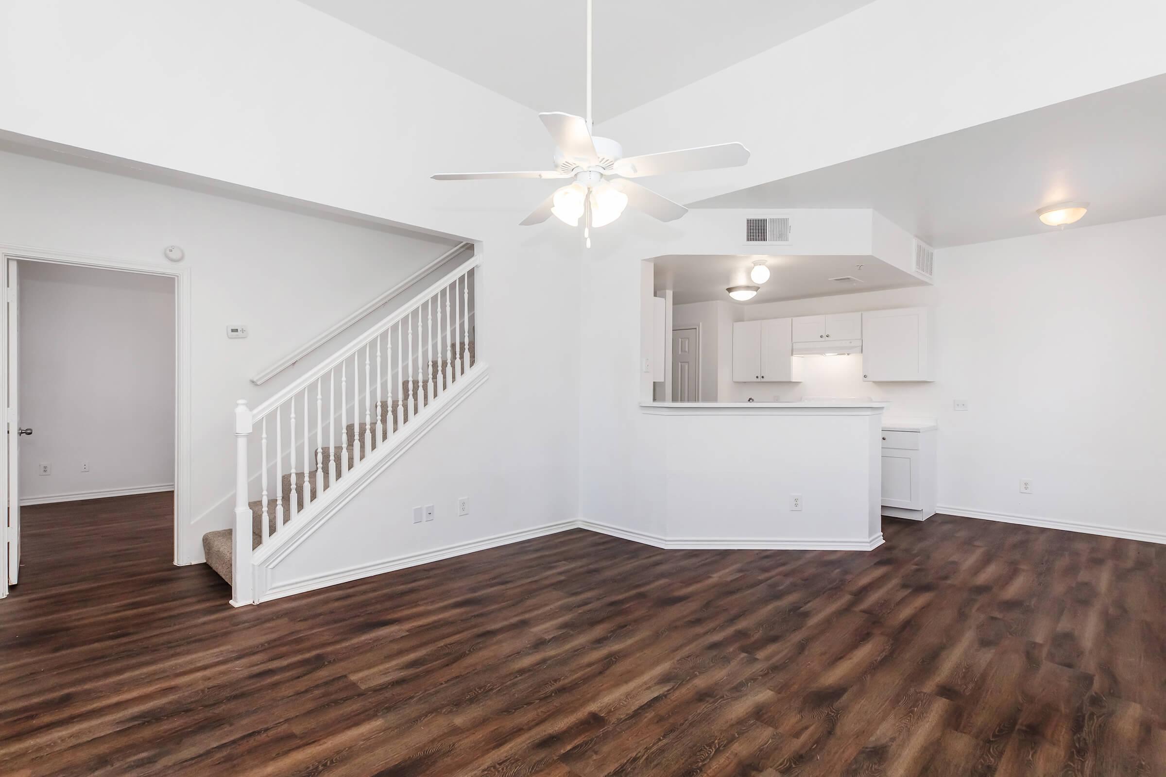 Spacious open-concept living area featuring a ceiling fan, laminate flooring, and a staircase leading to an upper level. The kitchen area includes white cabinets and a breakfast bar, with plenty of natural light illuminating the space. The walls are painted in a light, neutral color.