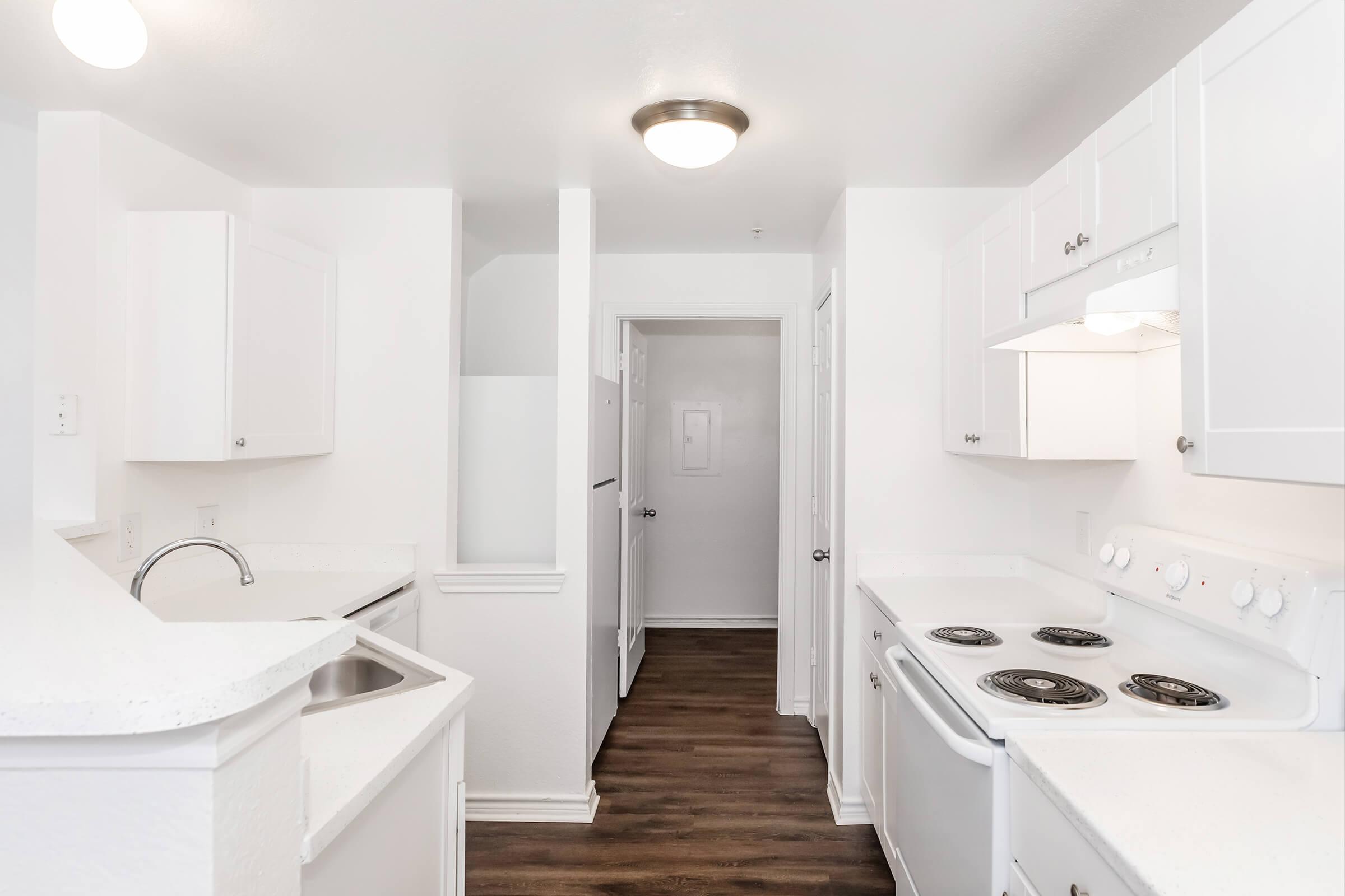 White kitchen featuring modern appliances, including a stove and refrigerator. The space has bright white cabinetry, a sink, and light hardwood floors. A hallway leads to an adjacent area, and the overall design is clean and contemporary. Natural light is present, enhancing the spacious feel.