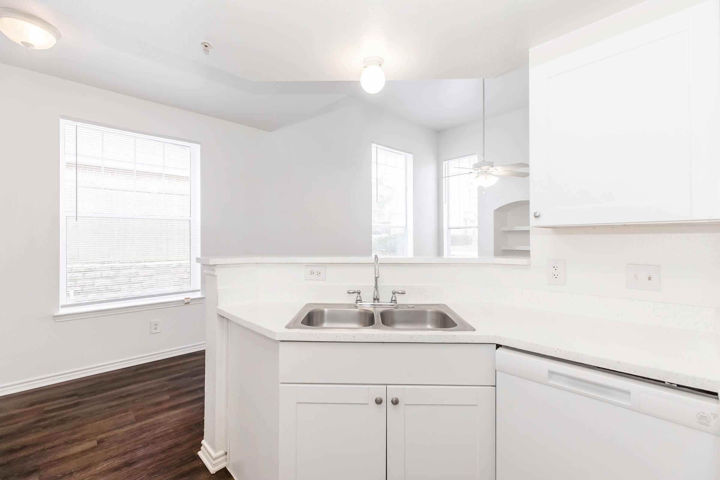 Clean and modern kitchen space featuring a double sink, white cabinetry, and a dishwasher. Natural light streams in through large windows, and the room has light brown hardwood flooring. The kitchen connects to a well-lit area, creating an open and inviting atmosphere.