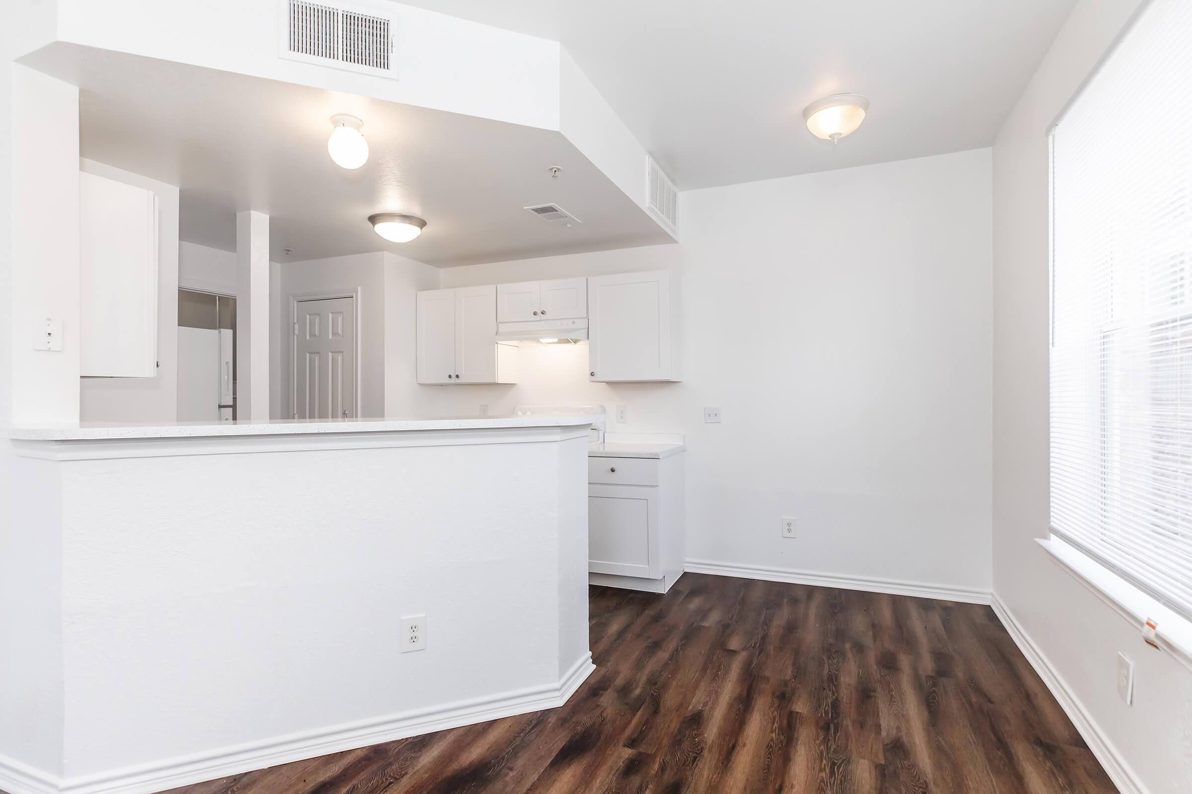 An interior view of a modern apartment featuring white walls and cabinets, a light-colored wood-like floor, and ample natural light coming through a window. The space includes a kitchen area with a small countertop and an open floor plan that connects to a living area.