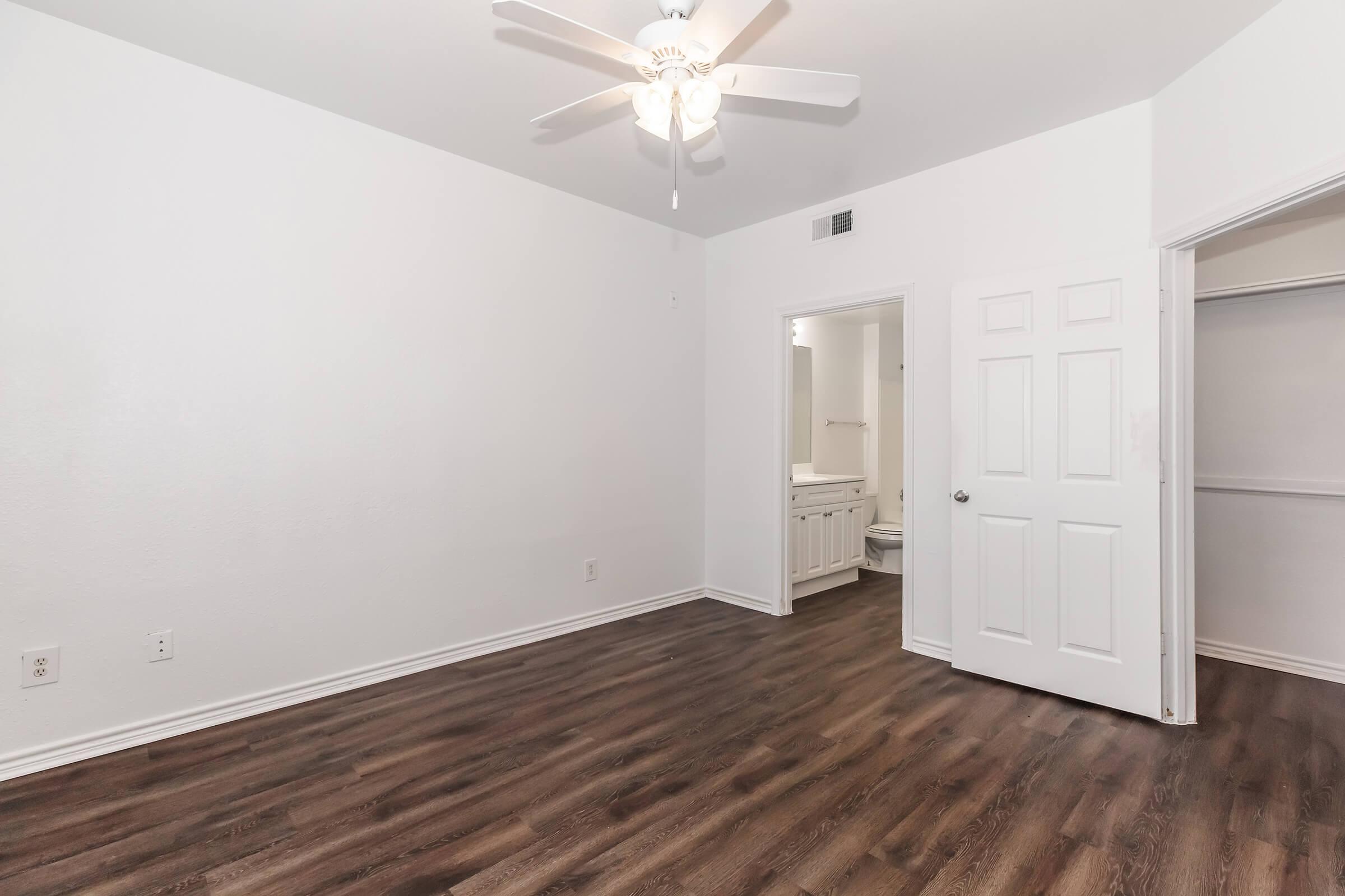 A spacious empty room with light-colored walls and a ceiling fan. There is an open doorway leading to a bathroom and another door that appears to lead to a closet. The floor is a dark wood laminate, providing a contrast to the walls. Natural light may be streaming in from a window.