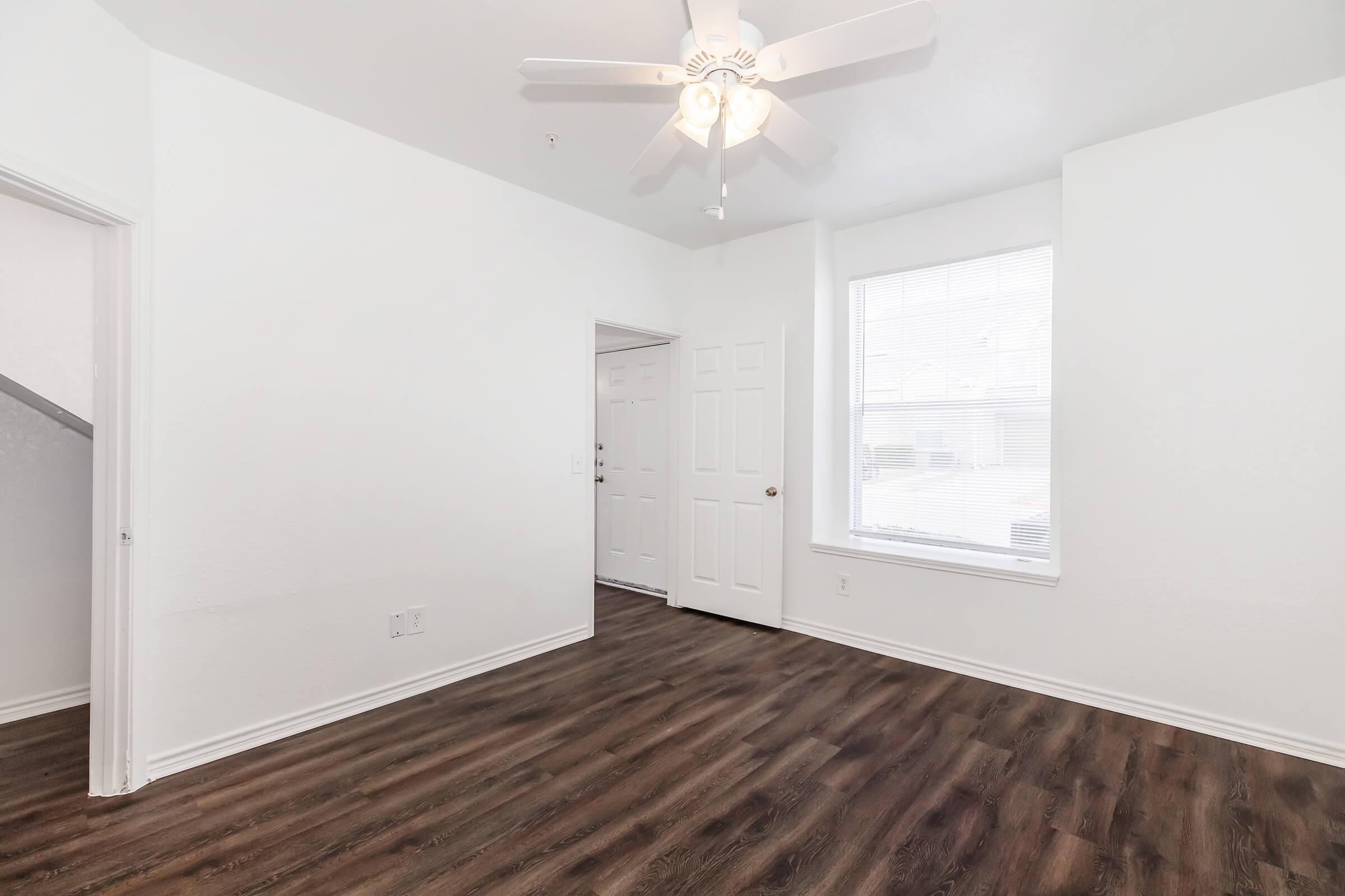 A bright, empty room with white walls and a ceiling fan. It has a window allowing natural light, wooden flooring, and a doorway leading to the entrance. The space is clean and minimalist, suitable for various interior design options.