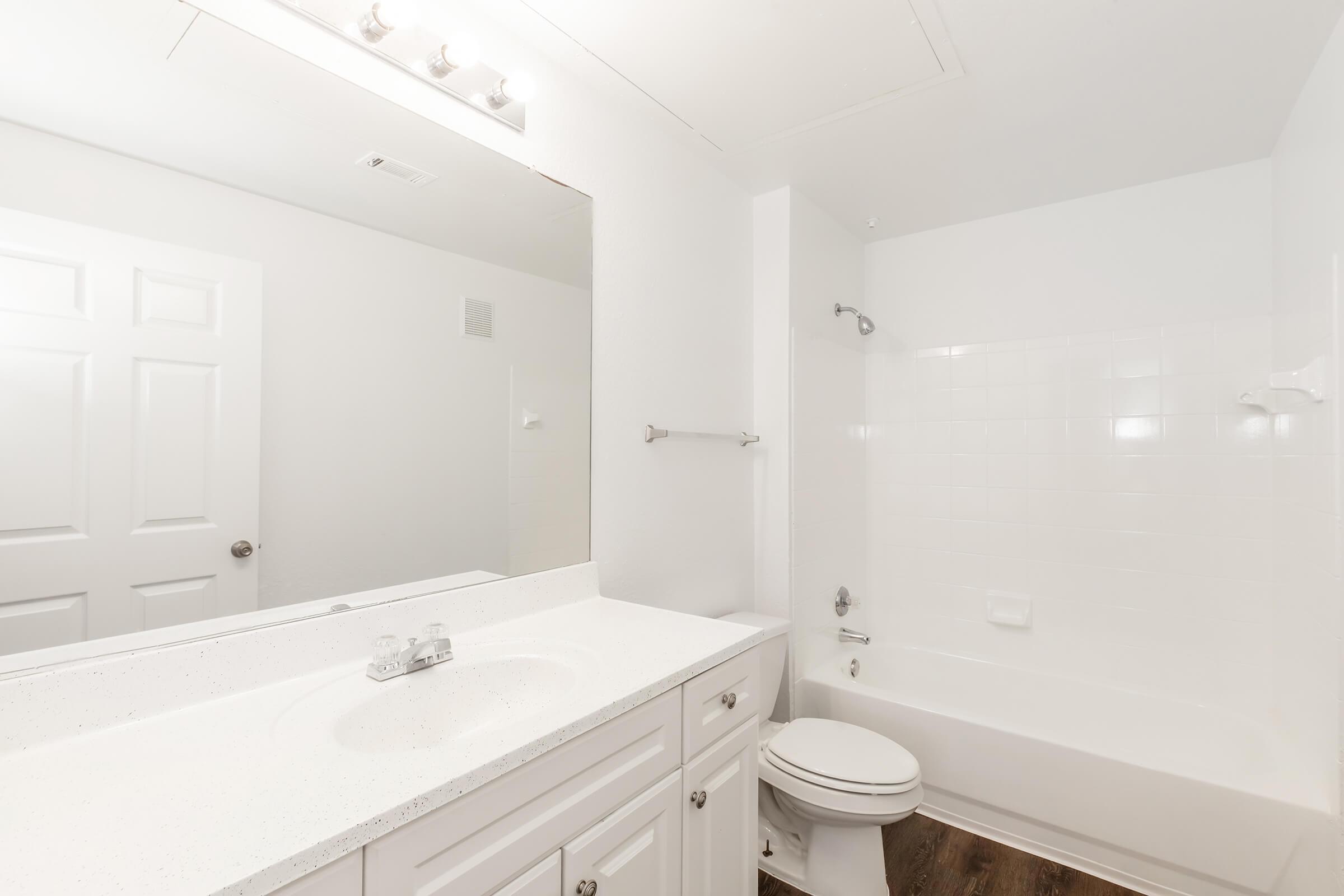 A bright, clean bathroom featuring a white sink with a modern countertop, a large mirror above, a white bathtub with a shower, and light-colored walls. The floor is dark, providing a nice contrast to the overall brightness of the space. A towel rack is mounted on the wall next to the bathtub.