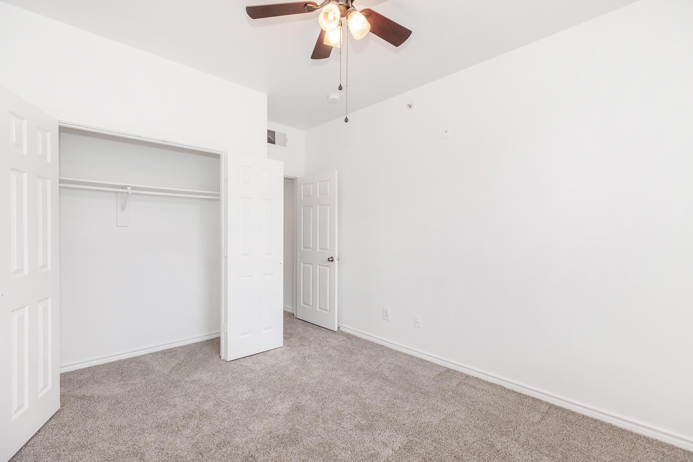 Empty room with light-colored walls and a ceiling fan. The floor is covered in beige carpet. There are two closed white doors, one leading to a closet. The room has no furniture, giving it a spacious and minimalist appearance.