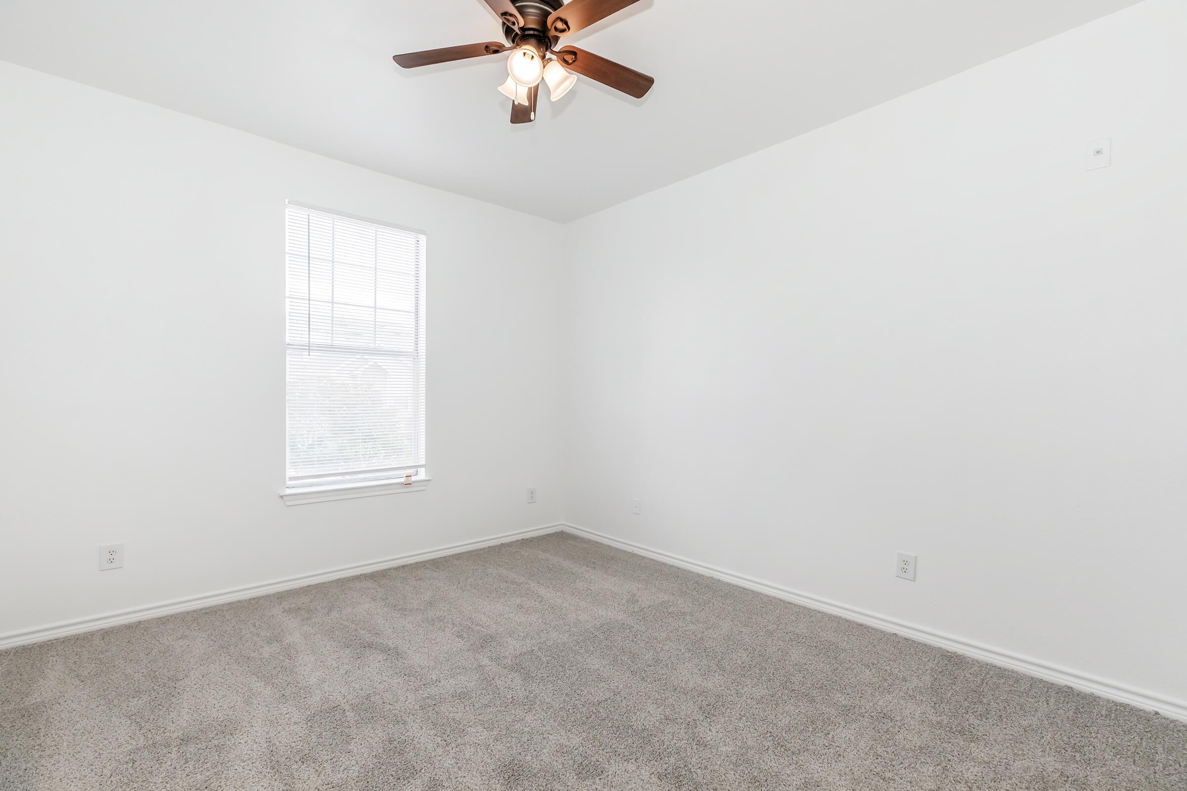 Empty room with light beige carpet and white walls. A ceiling fan with wooden blades is mounted in the center. There is a window with white blinds, allowing natural light to enter the space. The room is unfurnished, creating a blank canvas for potential interior design.