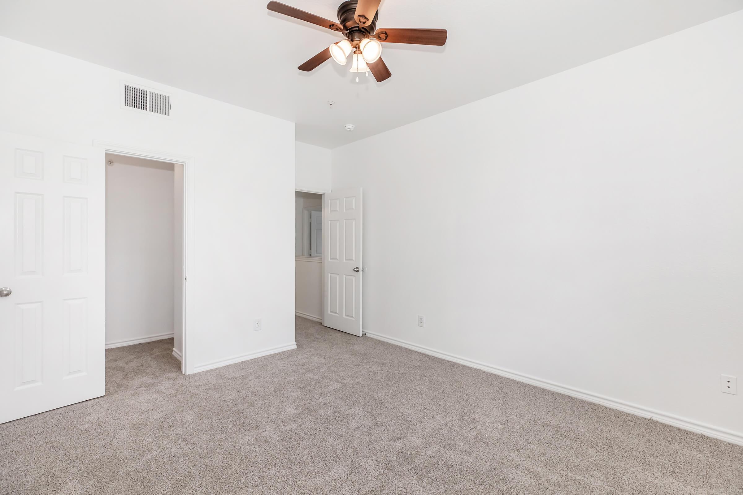 A bright, empty room with light-colored walls and a ceiling fan. The carpeted floor is a neutral shade. There are two doors; one leads to another area and the other is closed. The room is well-lit, creating a spacious and inviting atmosphere.
