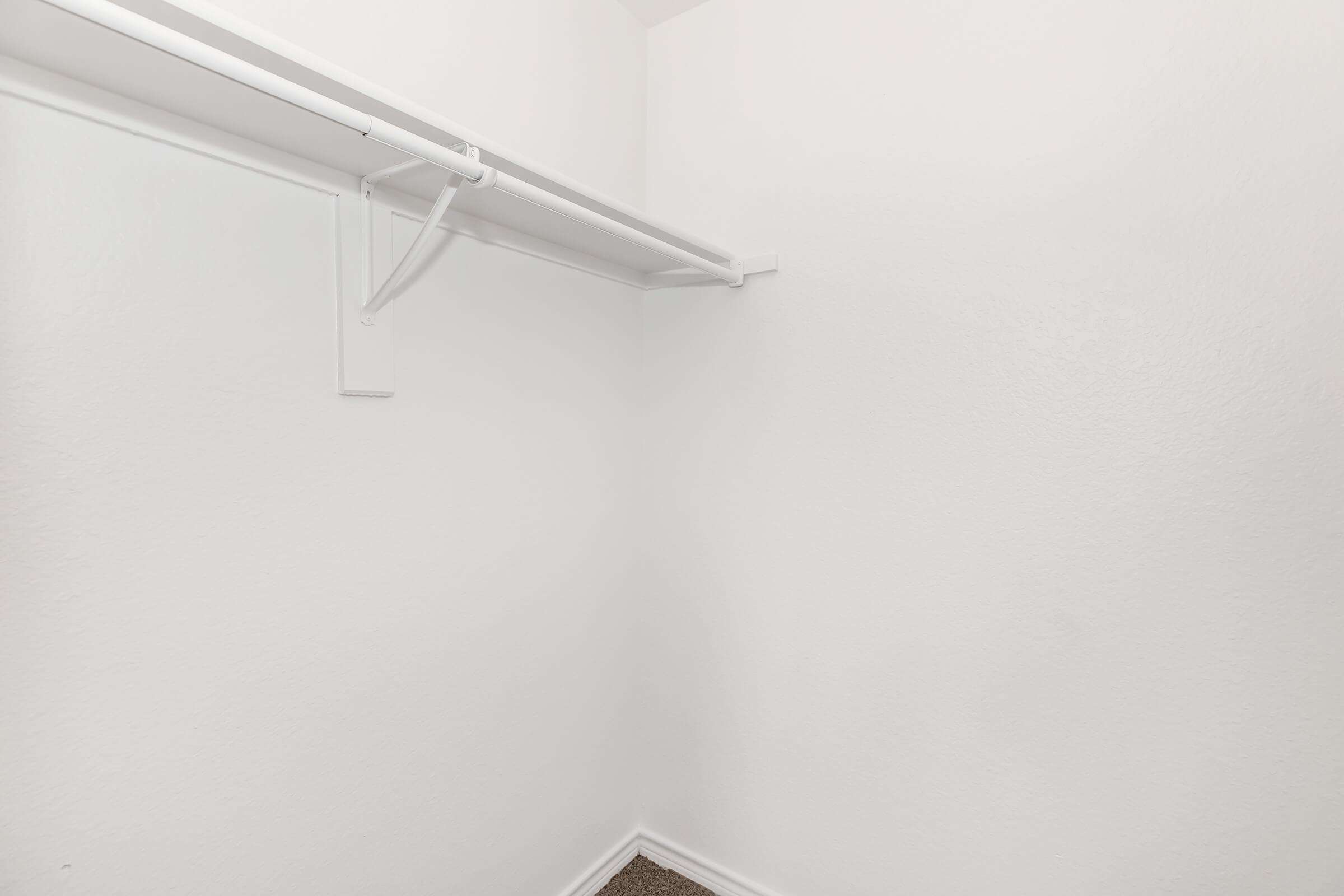 Empty closet with white walls and a single hanging rod along the top. The floor is covered in a brown carpet, and there are no visible items or storage solutions present. The space appears clean and minimalistic.