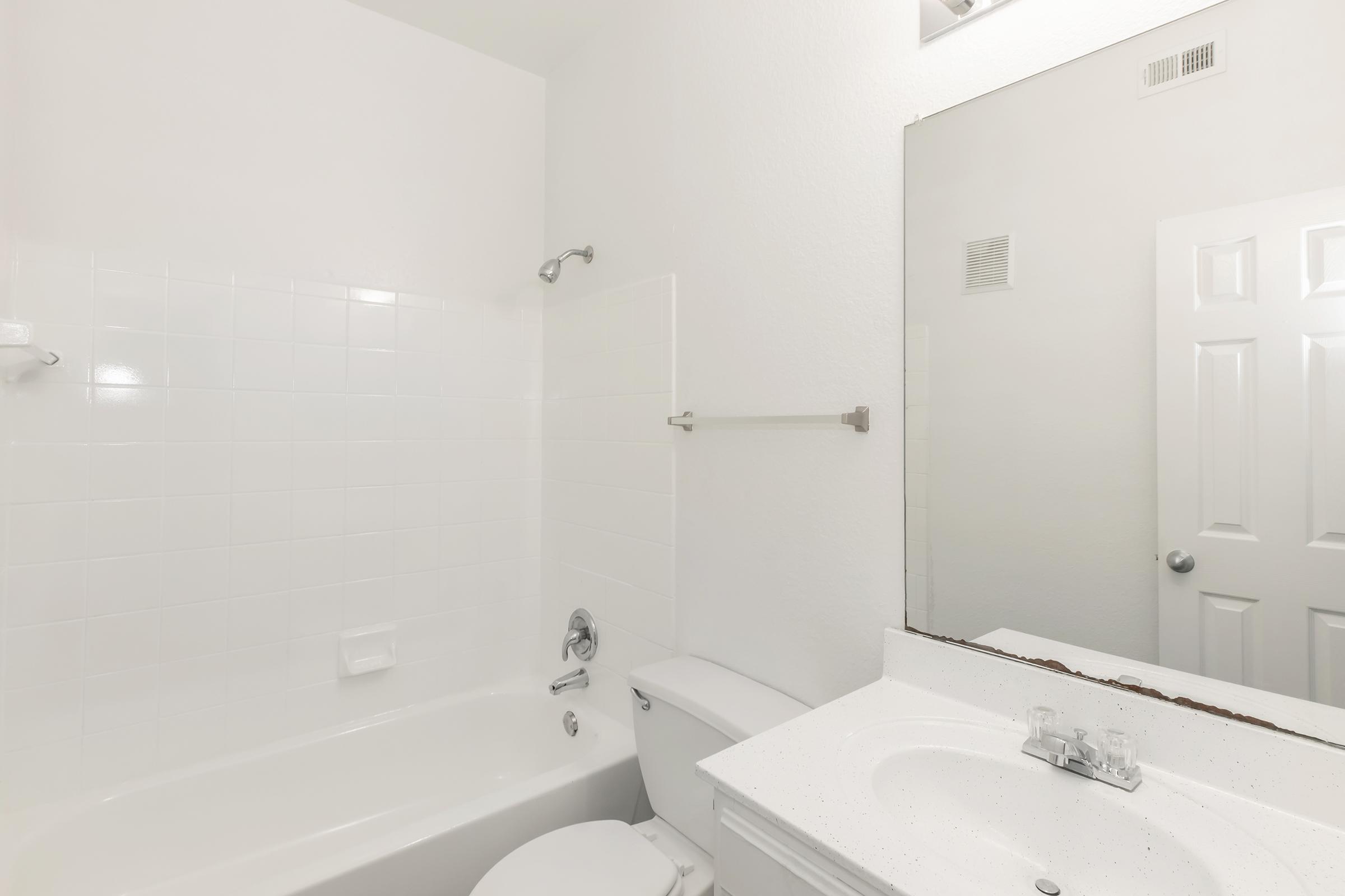 A clean, modern bathroom featuring white tile walls, a bathtub with shower, a white toilet, and a sink with a countertop mirror. The space is well-lit, with minimal decor, creating a fresh and tidy appearance. A door is visible in the background.