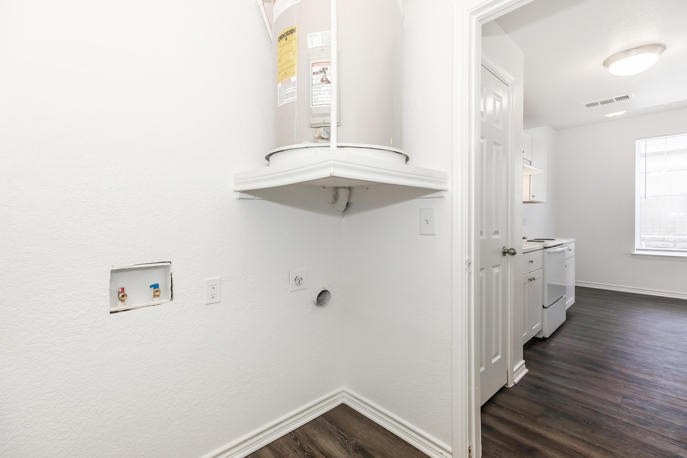 A small laundry area featuring a water heater mounted on the wall, with plumbing connections visible. The room has light-colored walls and dark wood-like flooring. A doorway leads to a kitchen area, and there is a vent outlet on the wall.