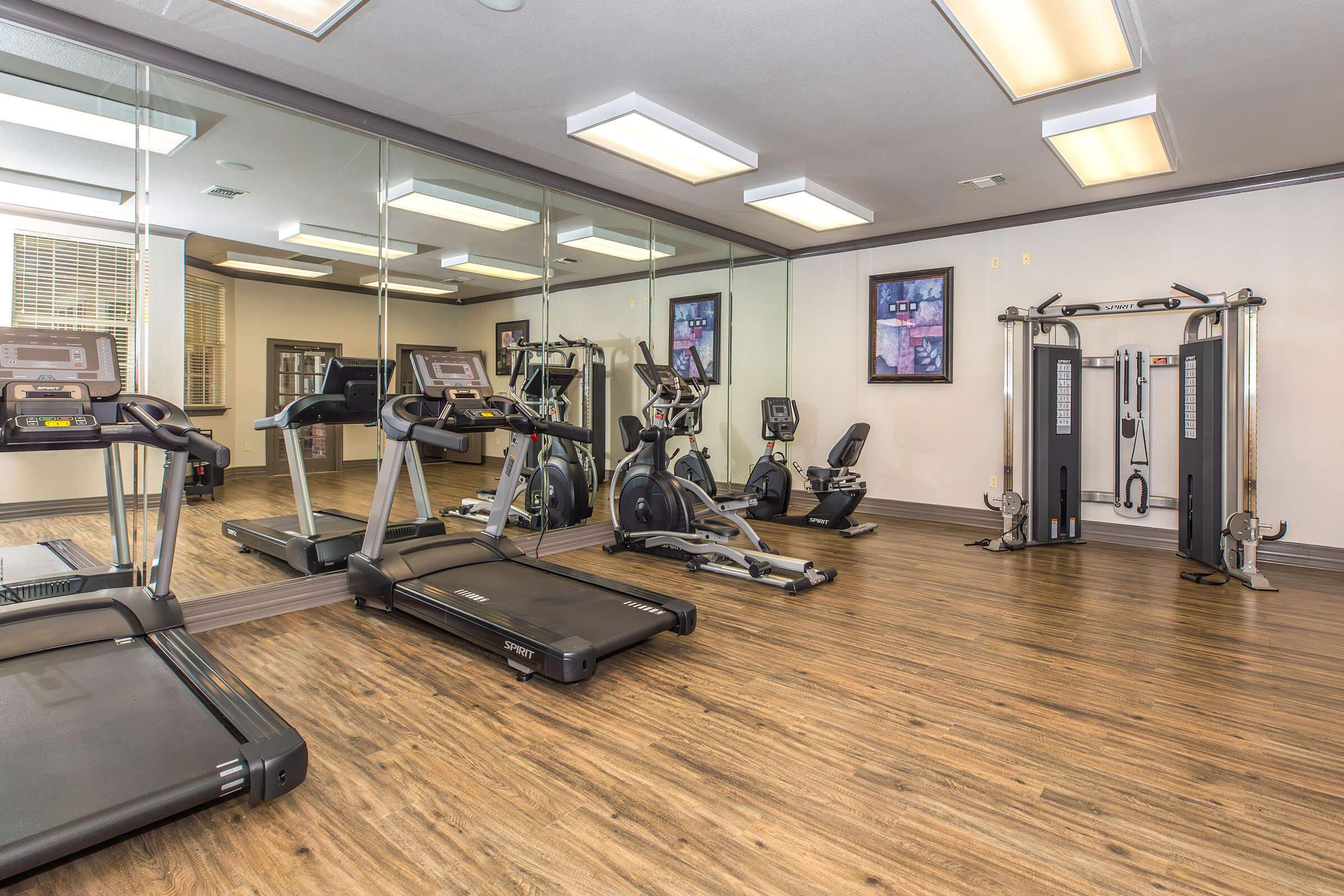 A modern gym interior featuring exercise equipment like treadmills, ellipticals, and strength training machines. The space is well-lit with mirrors on one wall and light wood flooring, creating a spacious and inviting atmosphere for fitness activities.