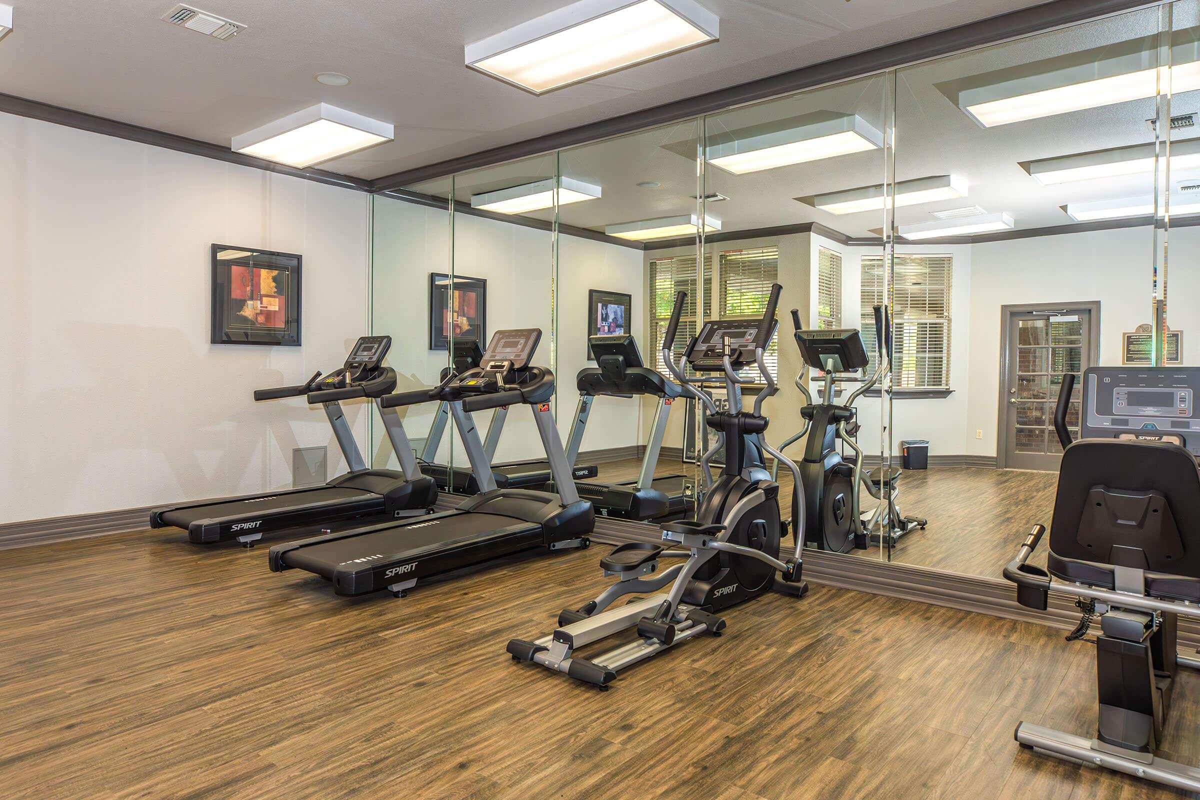 A modern fitness room featuring multiple cardio machines, including treadmills and exercise bikes. The walls are mirrored, enhancing the spacious feel. Simplistic decor includes framed artwork, and the room is well-lit with overhead lighting and windows allowing natural light.