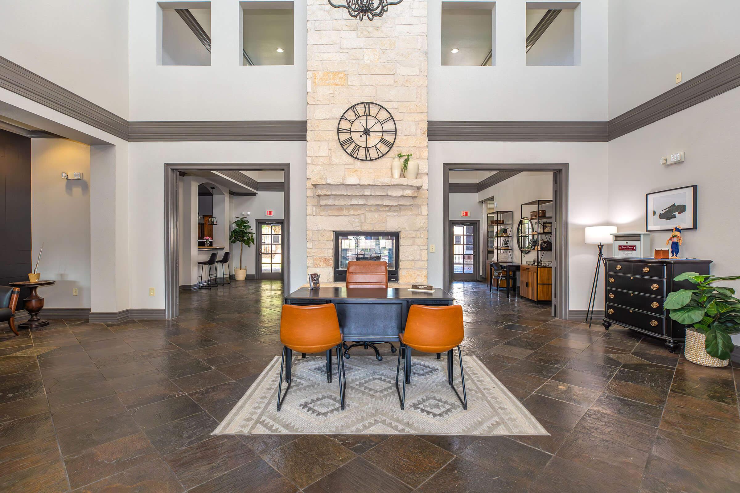 A spacious reception area featuring a large stone fireplace with a clock above it. In the center, there is a black desk with two brown chairs. The floor is tiled with dark stones, and a patterned rug lies under the desk. Light-colored walls and decorative plants add to the inviting atmosphere, with additional furniture visible in the background.