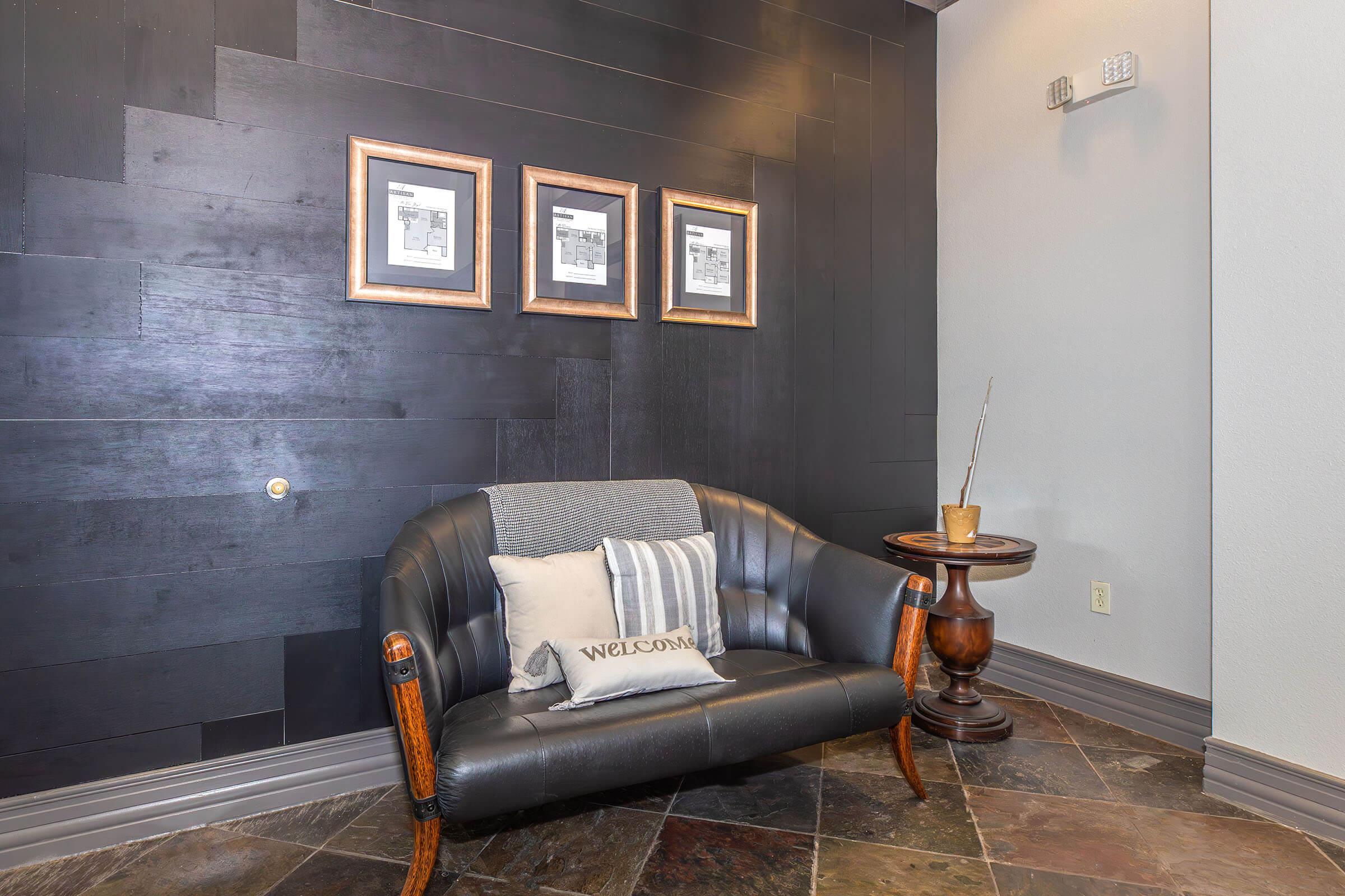 A cozy corner of a room featuring a black wall, a stylish two-seater sofa with a decorative pillow that says "WELCOME," a small wooden side table, and three framed black-and-white artworks. The floor is made of dark tiles, enhancing the modern aesthetic of the space.