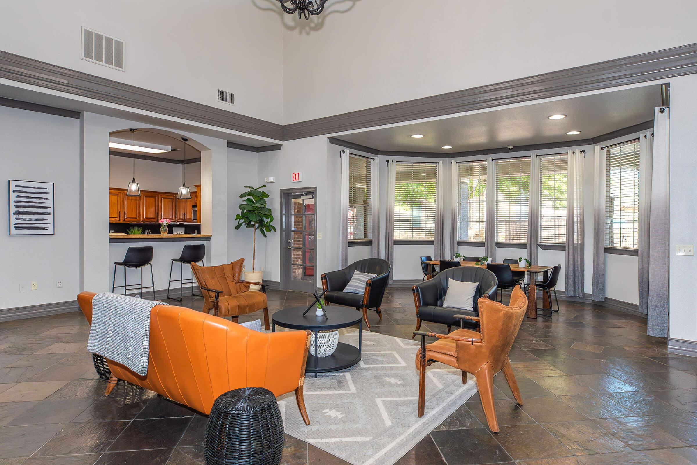 A modern lounge area featuring a mix of comfortable seating, including orange and black chairs, a round coffee table, and a patterned area rug. Large windows provide natural light, and the space includes plants and decor against neutral-colored walls, creating an inviting atmosphere.