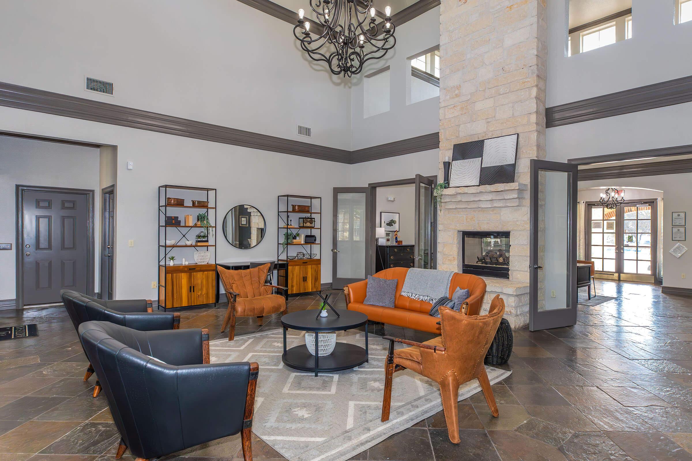 A spacious and modern living room featuring a large chandelier, stone fireplace, and stylish furniture. There are two brown leather armchairs, a round coffee table, and a gray area rug. Bookshelves and decorative elements add character, while large windows provide natural light. The walls are painted in neutral tones.