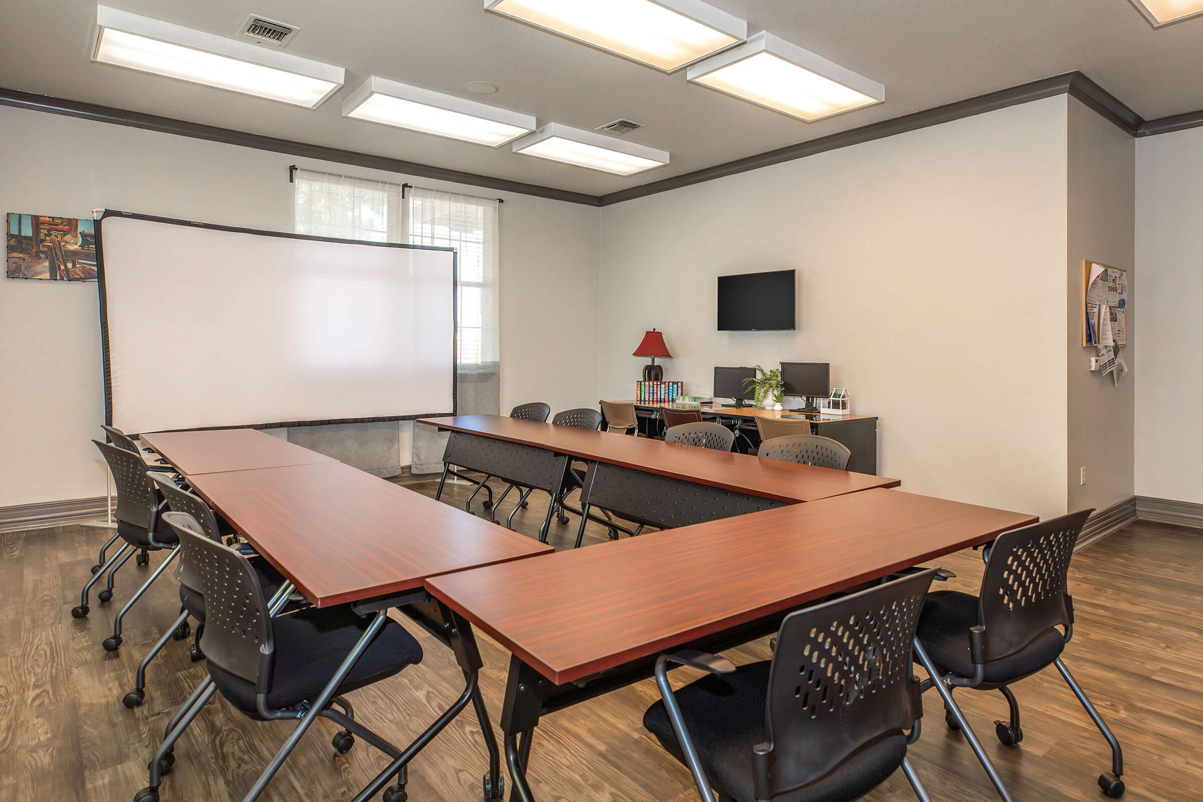 A modern conference room featuring multiple wooden tables arranged in a U-shape, surrounded by black office chairs. A large screen for presentations is positioned at the front, with natural light coming through a window. The room has a TV, a small lamp, and decorative wall art.