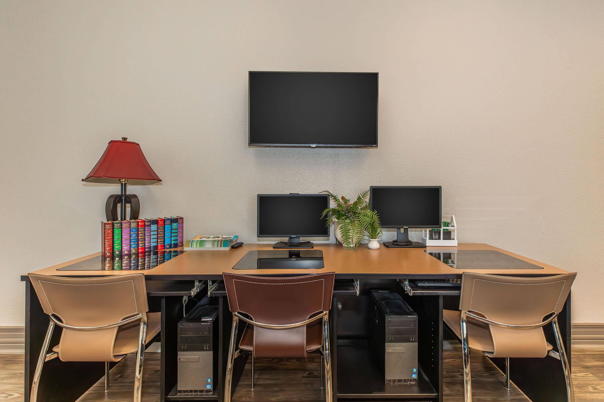 A modern home office setup featuring two computer monitors on a wooden desk, a potted plant, and a stack of colorful books. A flat-screen TV is mounted on the wall above the desk. Three stylish chairs are placed around the desk, with two computer towers positioned under the desk. A lamp with a red shade adds a touch of warmth.