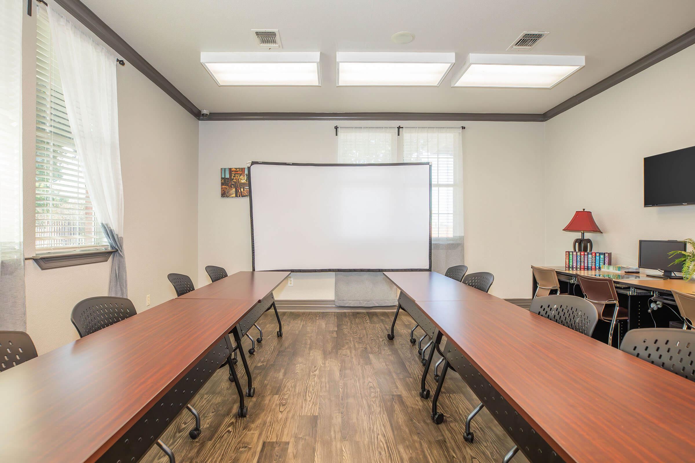 A spacious meeting room featuring two long tables arranged in a U-shape. A projector screen is positioned at the front, with a red lamp and shelves with books on one side. The room has large windows letting in natural light and is decorated with a few pieces of artwork.