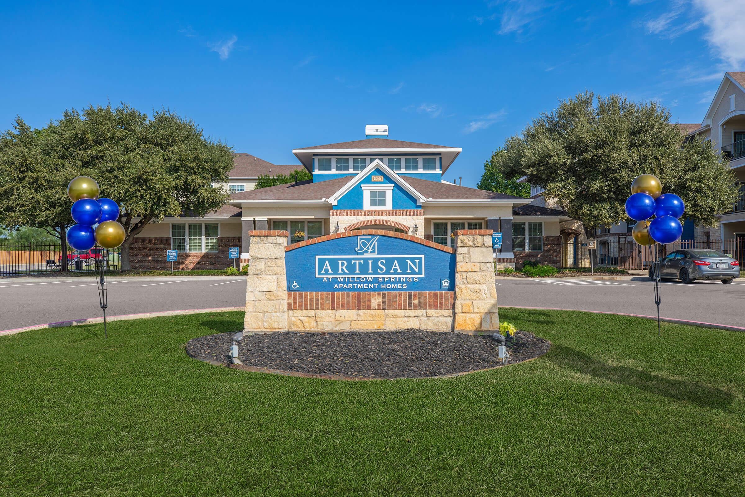 Sign for Artisan Apartments featuring a stone base with the name "ARTISAN" prominently displayed. Flanking the sign are decorative balloons in blue and gold. The entrance is set in a landscaped area with green grass, surrounded by trees and a residential building in the background.