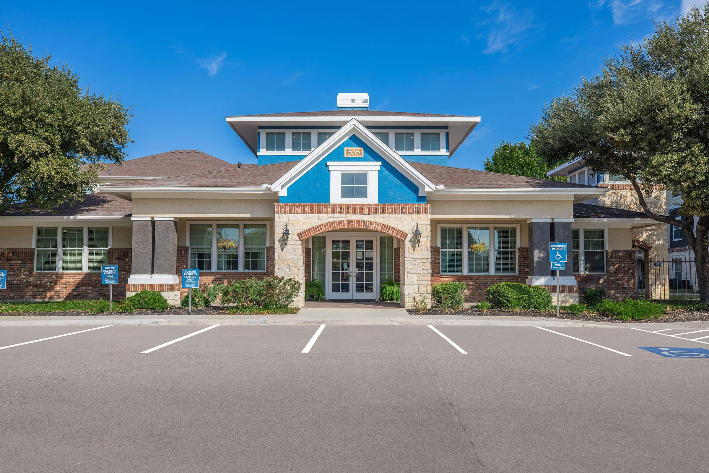 A modern residential office building featuring a brick and blue exterior, surrounded by trees and manicured bushes. The entrance has large windows and a welcoming porch, with several parking spaces visible in front. Clear skies and bright lighting enhance the overall appearance.