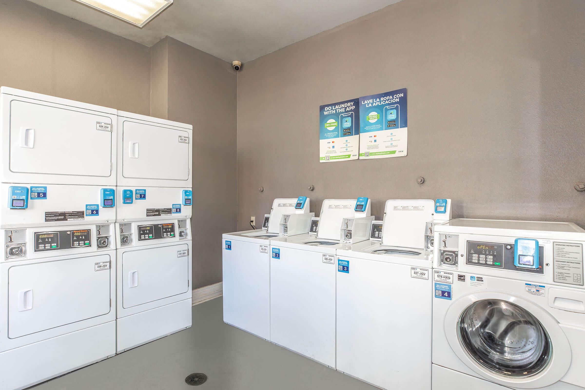 A laundry room featuring multiple commercial washing machines and dryers. The machines are white with blue interface panels. A wall-mounted sign above provides instructions for use. The room has gray walls and a ceiling light.