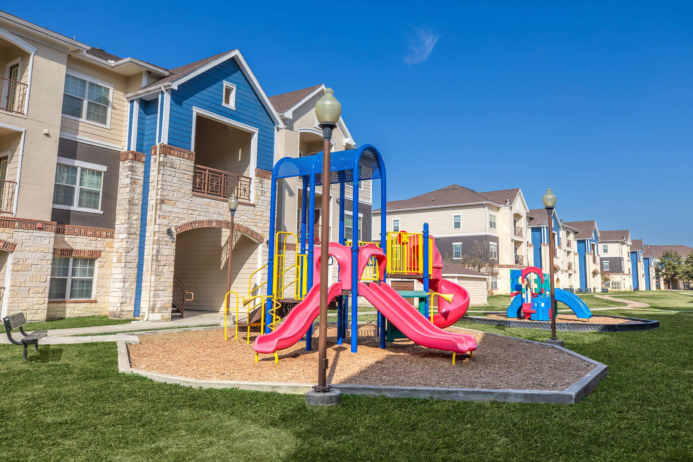 Colorful playground equipment with slides and climbing structures sits on a bed of wood chips in a grassy area. Surrounding the playground are residential apartment buildings with balconies. The sky is clear and blue, creating a bright and inviting atmosphere.