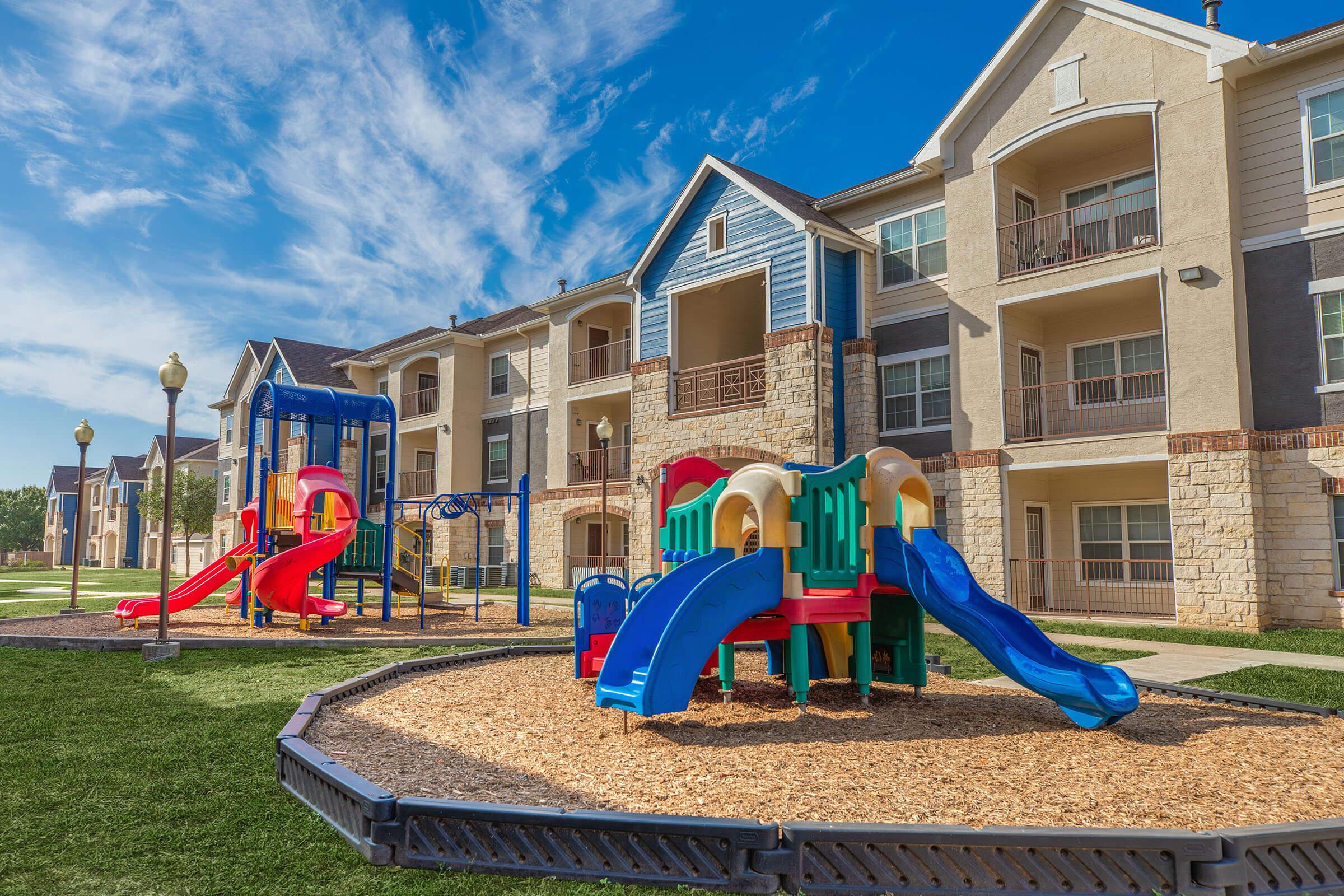 Colorful playground equipment featuring slides and climbing structures, set against the backdrop of modern apartment buildings. The ground is covered in wood chips, and the scene is bathed in bright sunlight with a clear blue sky. It's a vibrant space designed for children's play and recreation.