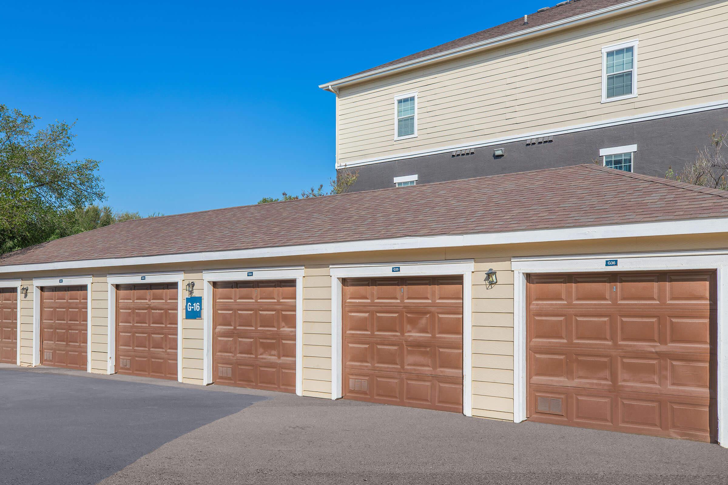 A row of closed garage doors with brown panels, set against a clear blue sky. The garage structure features a sloped roof and is part of a larger building visible in the background, which has beige and dark gray siding. The scene is well-lit, conveying a tidy and organized appearance.