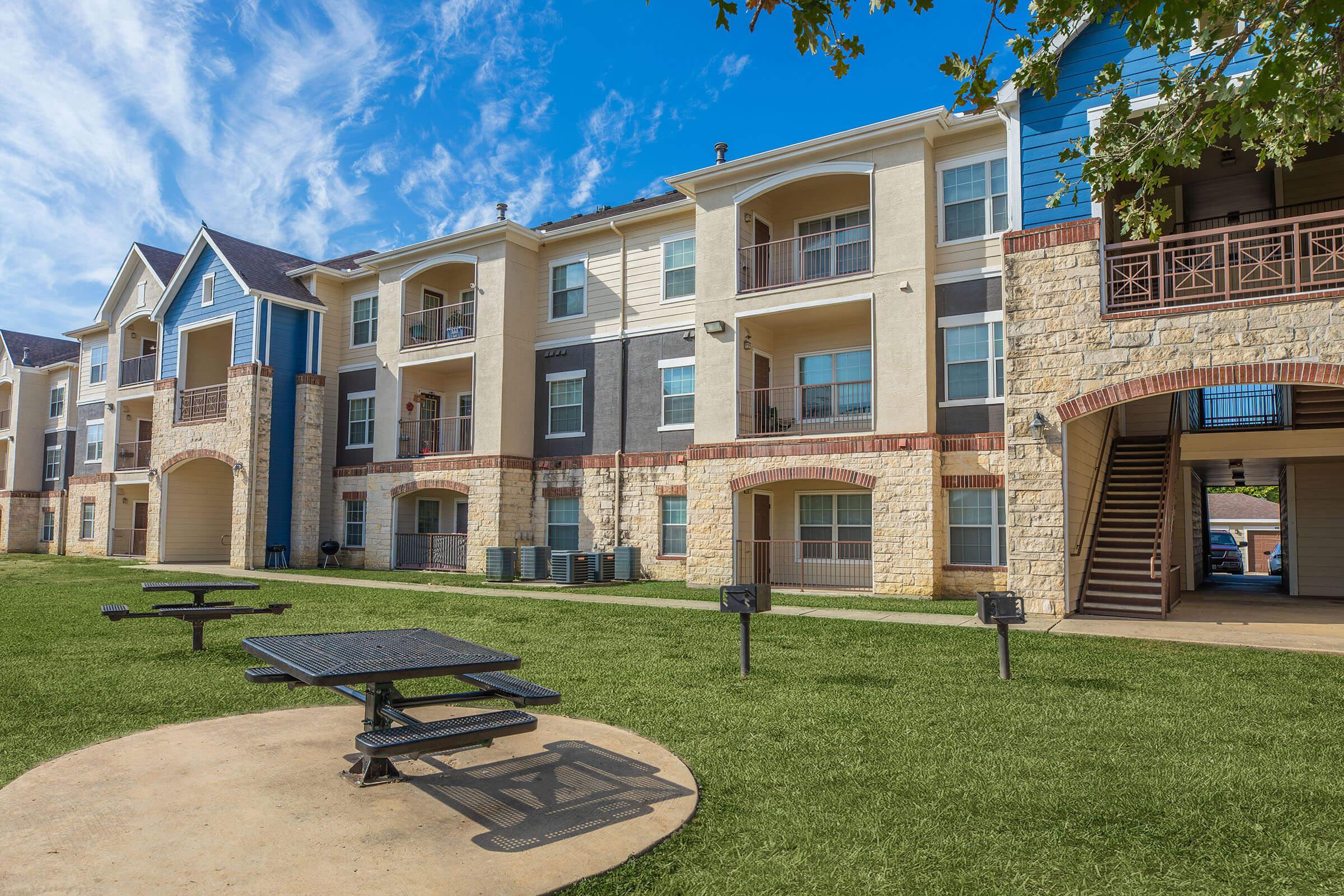 A view of an apartment community featuring multiple buildings with a mix of blue and beige exteriors. In the foreground, there is a circular picnic table on a grassy lawn, with outdoor grills visible nearby. The sky is clear and bright, suggesting a sunny day.