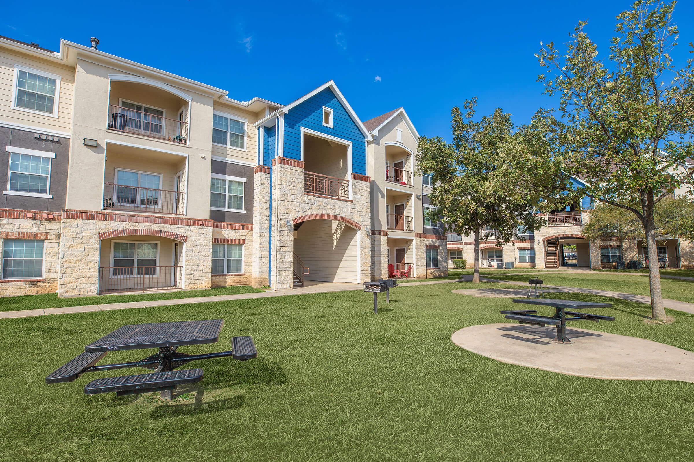 A well-maintained apartment community with a green lawn, picnic tables, and trees under a clear blue sky. The building features a mix of light-colored and blue siding, with balconies visible on the upper levels.