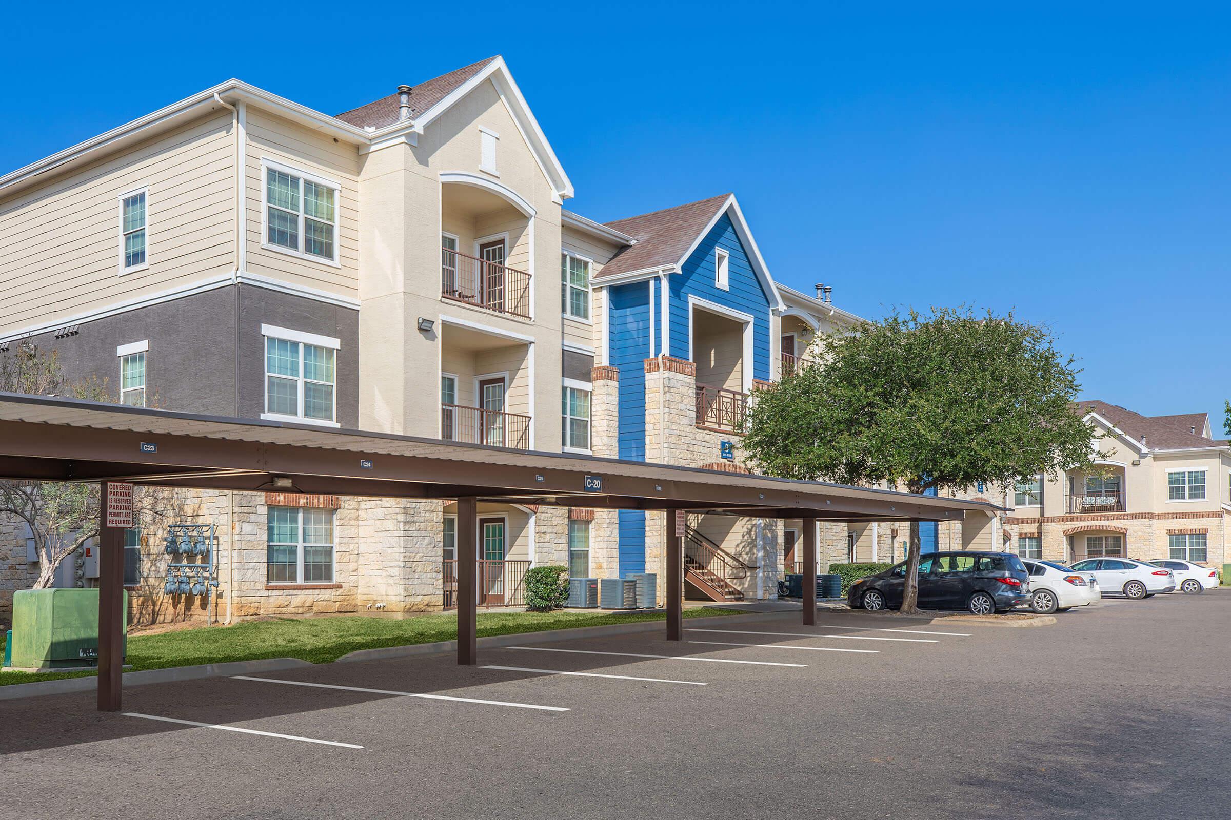 Exterior view of a residential apartment community featuring multi-story buildings with a mix of siding colors, including beige, gray, and blue. There is a covered parking area in the foreground with empty parking spaces, and lush green landscaping nearby under a clear blue sky.