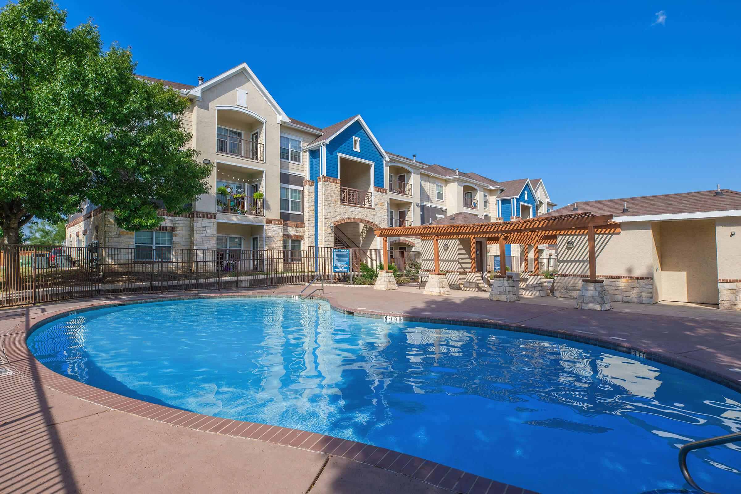 A bright and sunny view of an apartment community featuring a sparkling swimming pool surrounded by stone pillars and wooden pergolas. The buildings display a mix of beige and blue exteriors, with greenery from nearby trees and a clear blue sky in the background, creating a welcoming outdoor atmosphere.