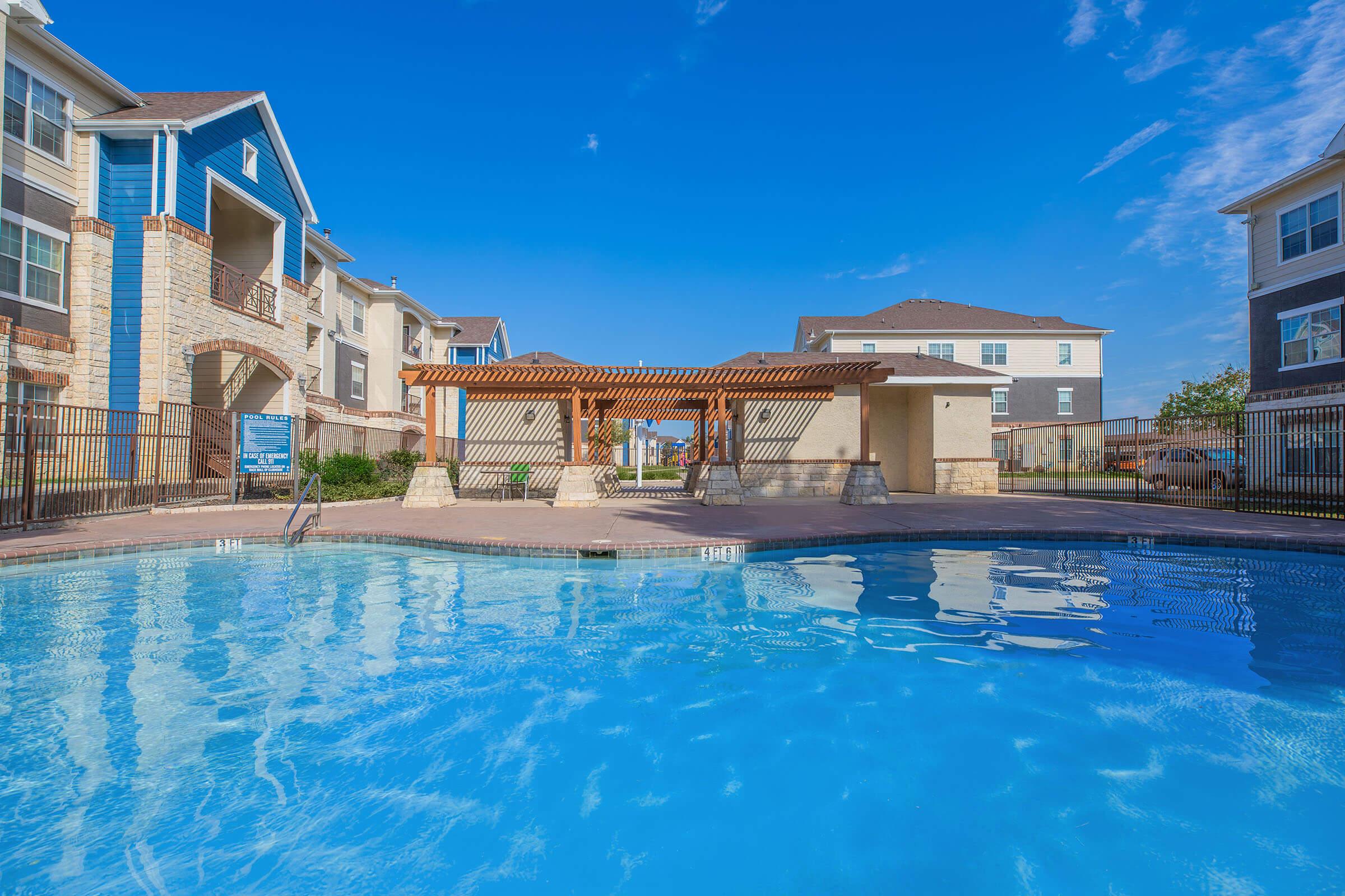 A clear blue swimming pool surrounded by a fenced area, featuring a wooden pergola and neatly landscaped grounds. Modern apartment buildings in the background under a bright blue sky with a few clouds. The scene conveys a relaxing outdoor atmosphere.