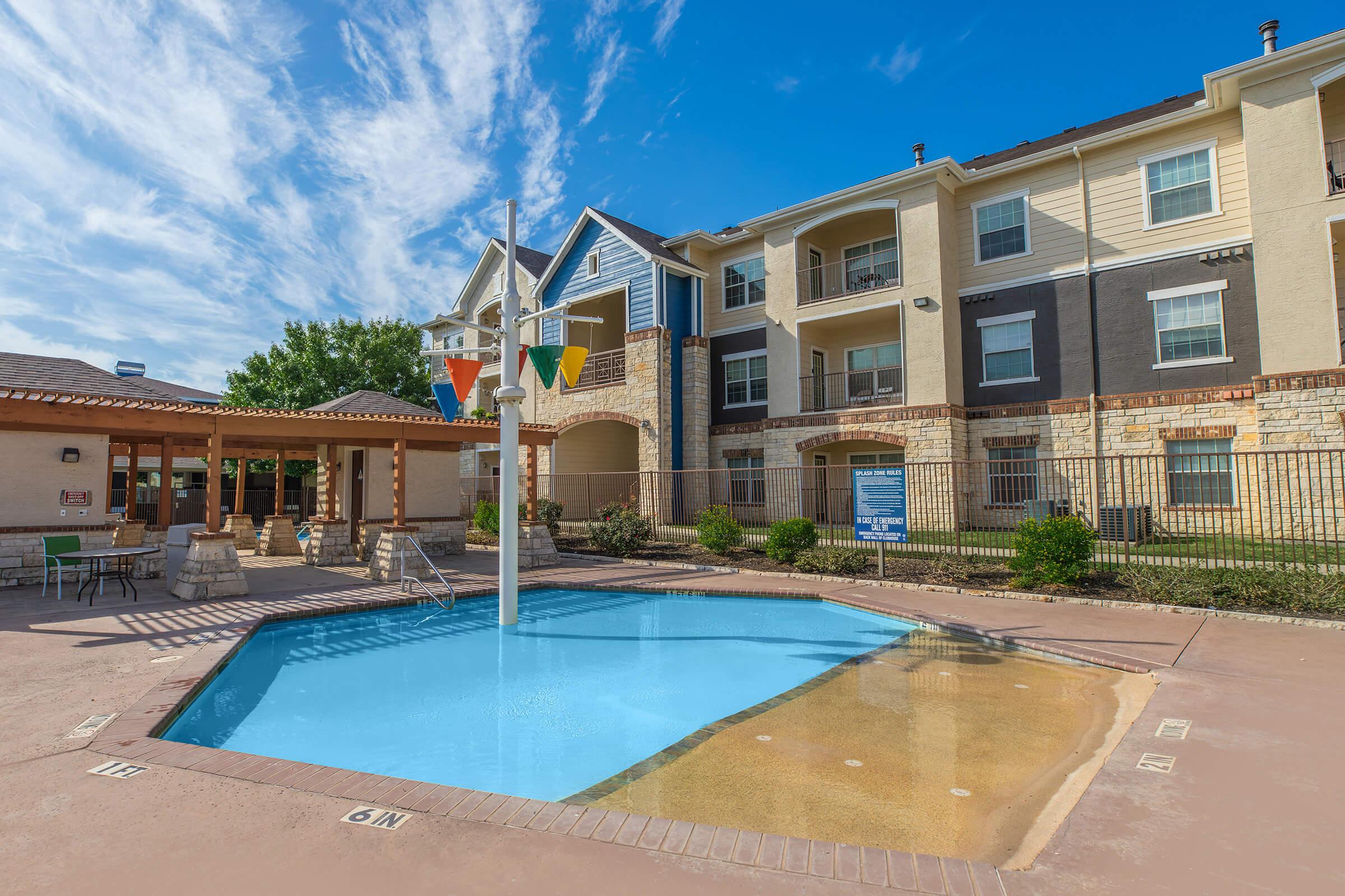 Swimming pool with a colorful water play structure, surrounded by a patio area and lush landscaping. Apartment building in the background features a mix of stone and wood siding, with blue skies and a few clouds above. The area is well-maintained and inviting for residents.