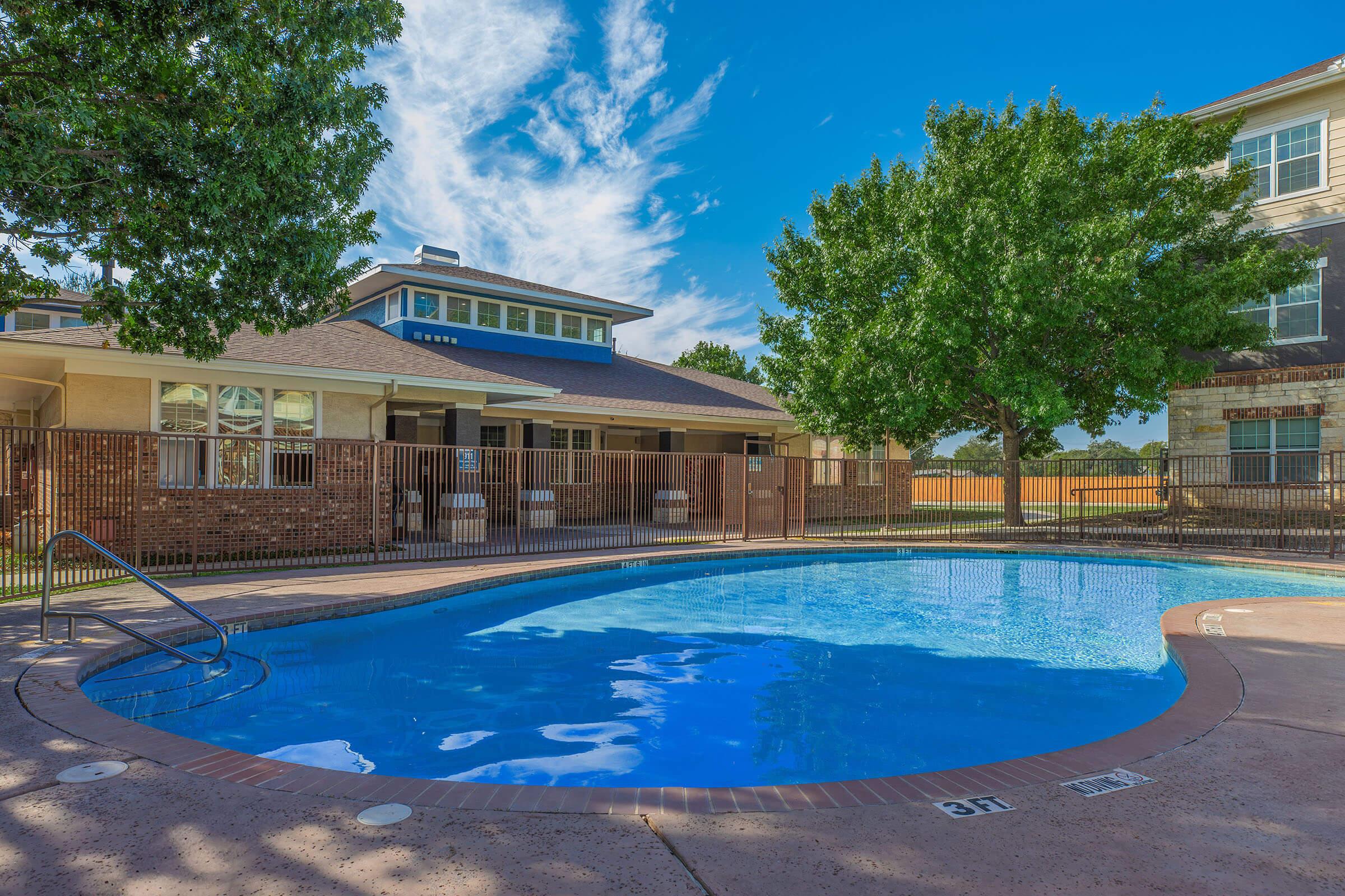 A clear swimming pool surrounded by a safety fence, with a ladder on one side. Lush green trees are visible nearby, and a two-story building is in the background under a bright blue sky with scattered clouds. The setting is well-maintained and inviting.