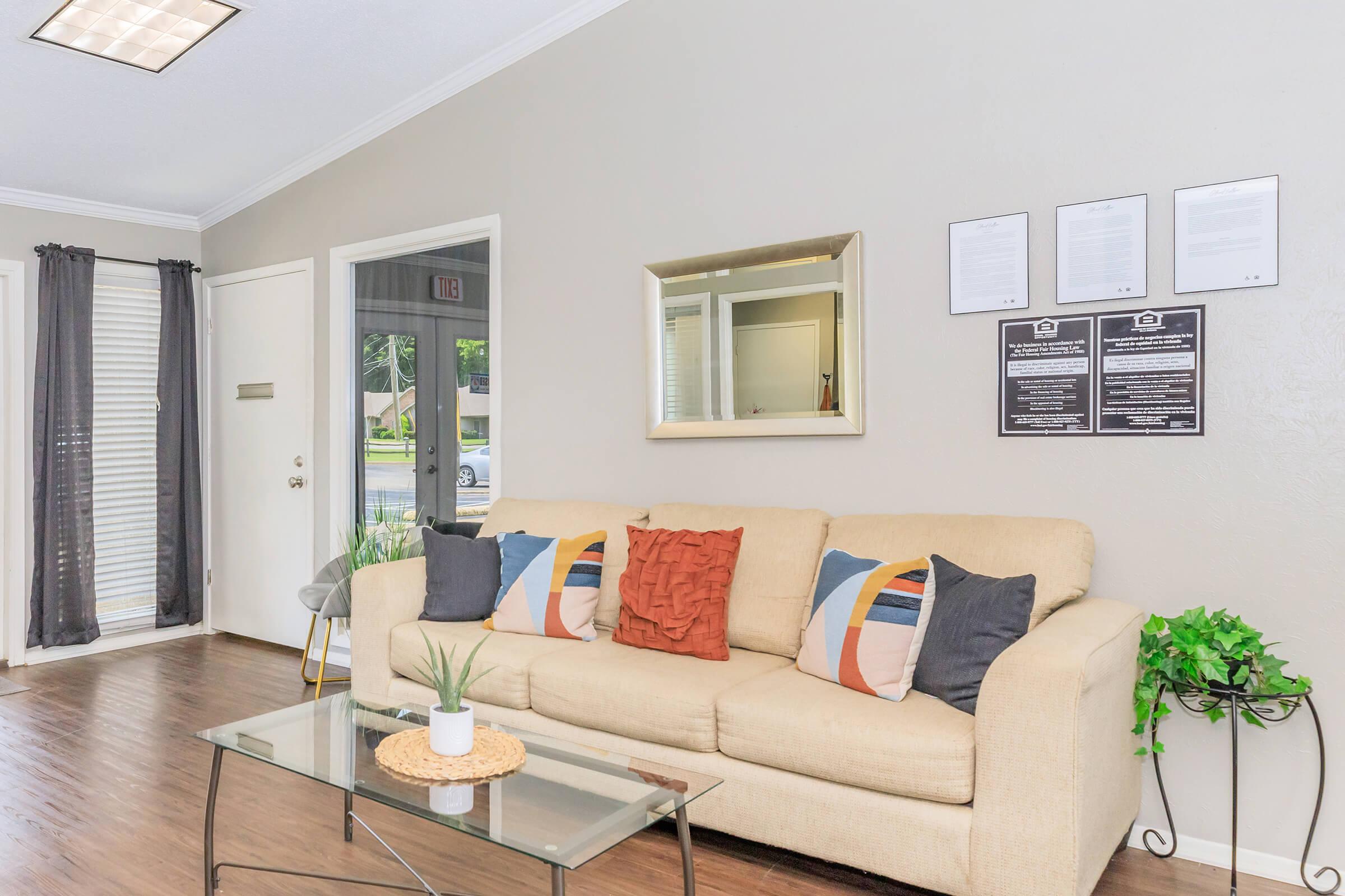 A cozy living room featuring a light-colored sofa with colorful pillows, a glass coffee table, and a small potted plant. There are two windows with sheer curtains, a mirror on the wall, and framed documents neatly displayed. The floor is dark wood, contributing to a warm ambiance.