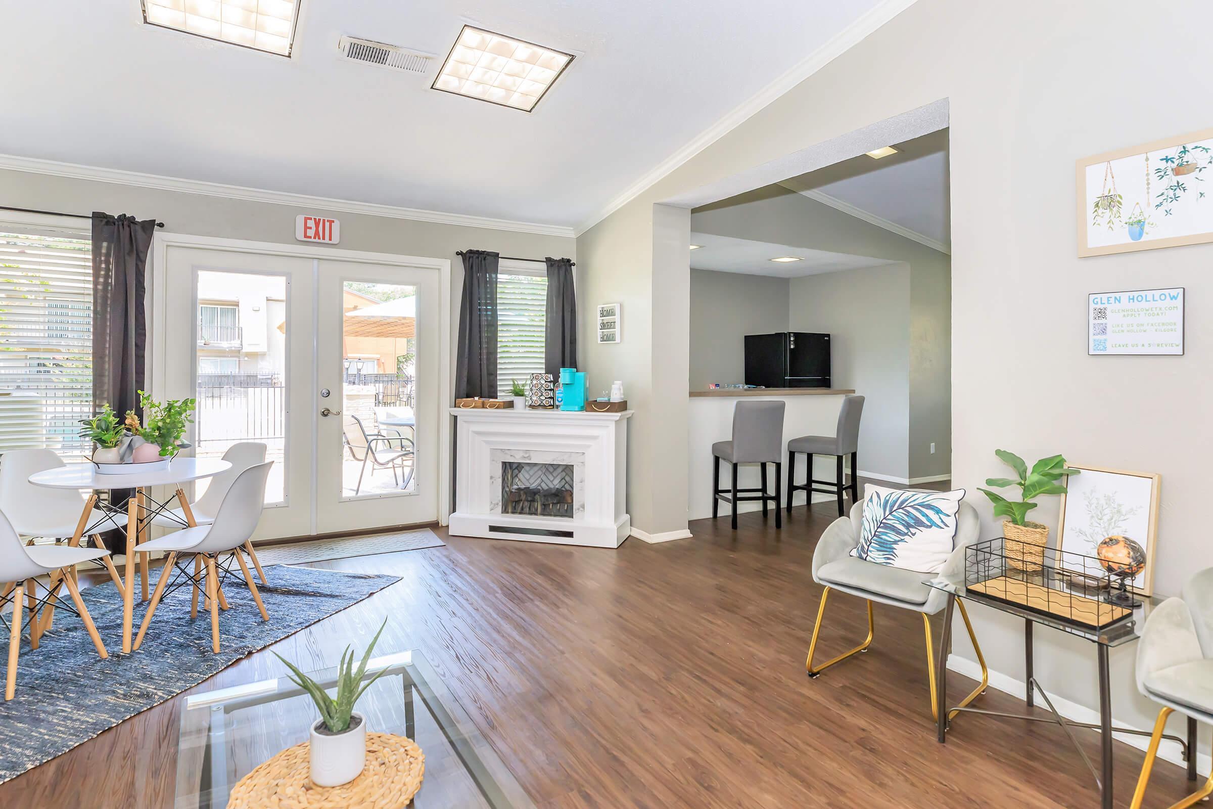 Living room area featuring a dining table with white chairs, a cozy seating arrangement, and a fireplace. Large windows with curtains allow natural light. In the background, there’s a bar area with stools and a view of an outdoor space. The decor is modern and inviting, with green plants adding a touch of nature.