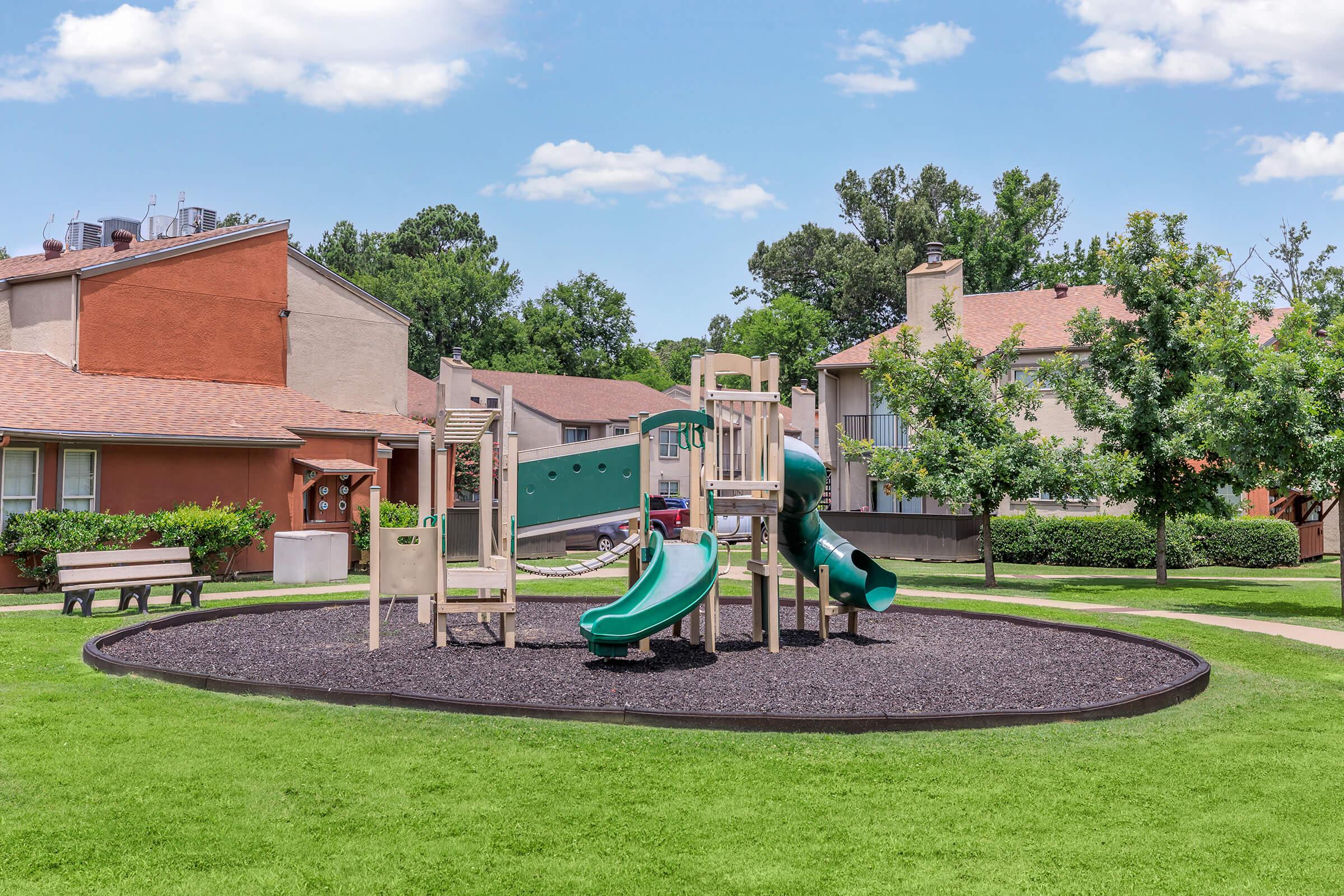 Playground with a wooden play structure featuring slides and climbing areas, set on a circular area of black mulch. The playground is surrounded by green grass and trees, with a few residential buildings in the background. Bright blue sky with a few clouds overhead.