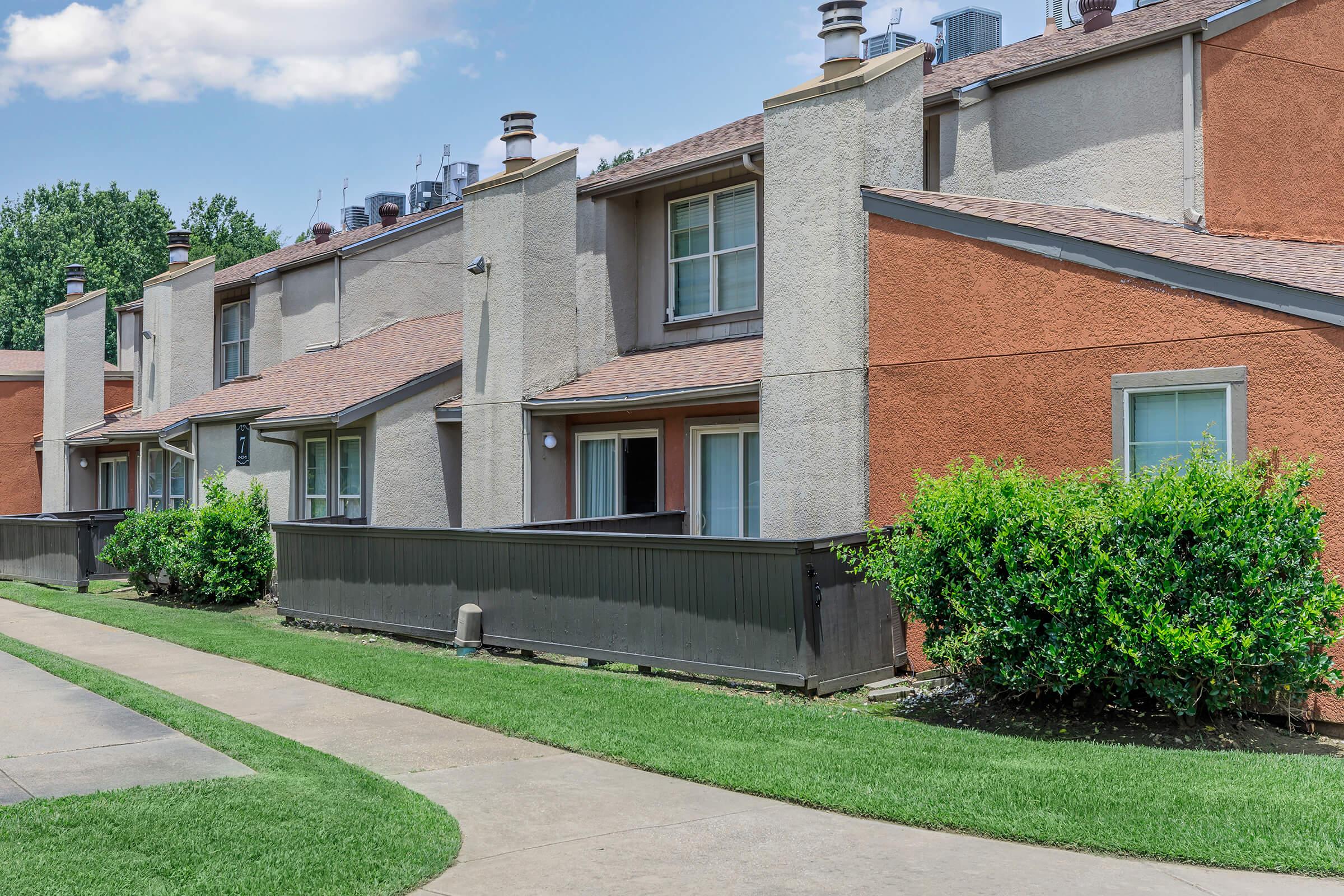 A row of multi-story residential buildings with a mix of beige and orange exteriors. Each unit features a porch and large windows. A well-maintained lawn and pathway are visible in the foreground, with trees and shrubs lining the area, under a clear blue sky.