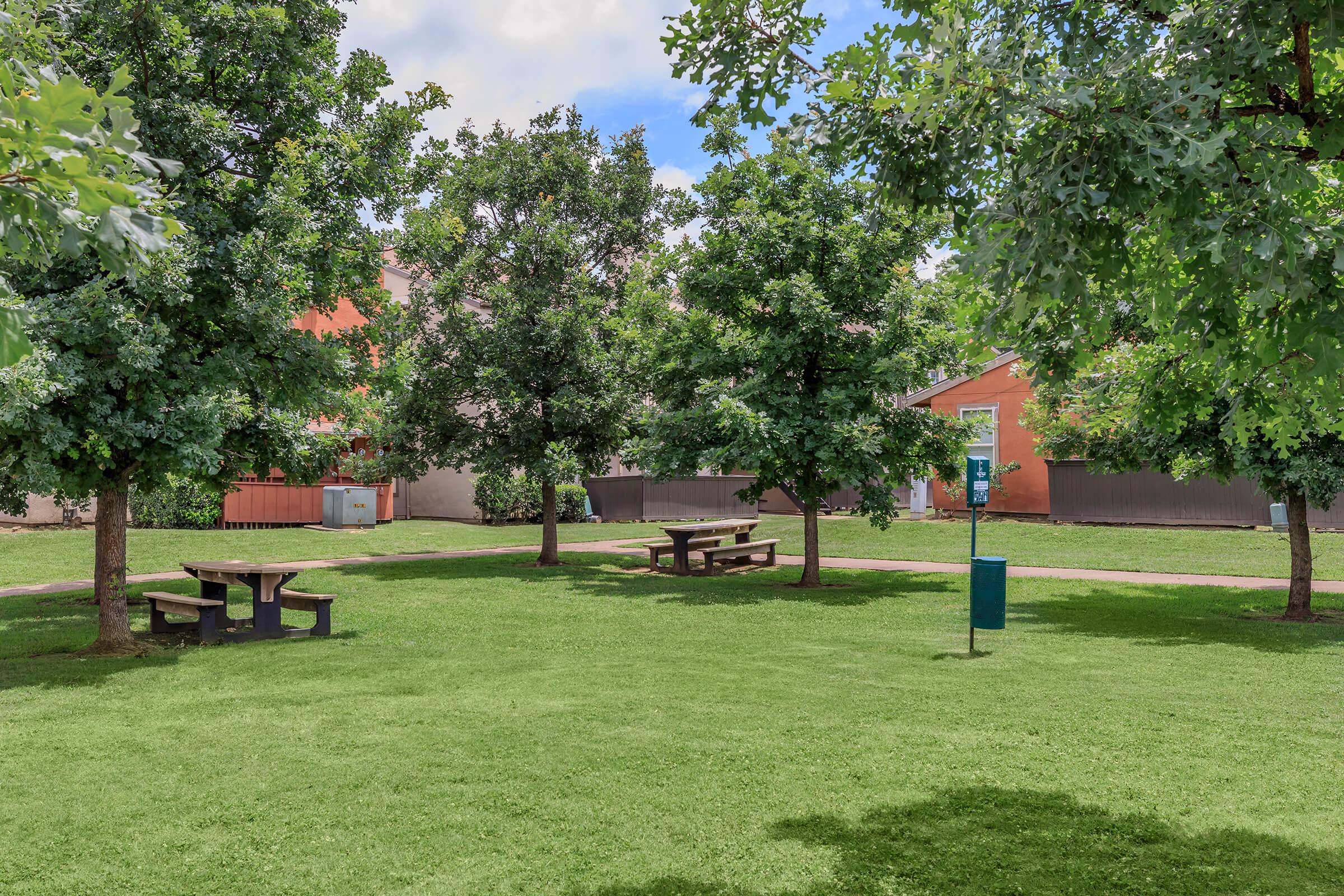 A grassy park area featuring several green trees and wooden picnic tables. In the background, there are some buildings and a trash bin. The sky is partly cloudy, creating a serene outdoor setting suitable for relaxation or gatherings.