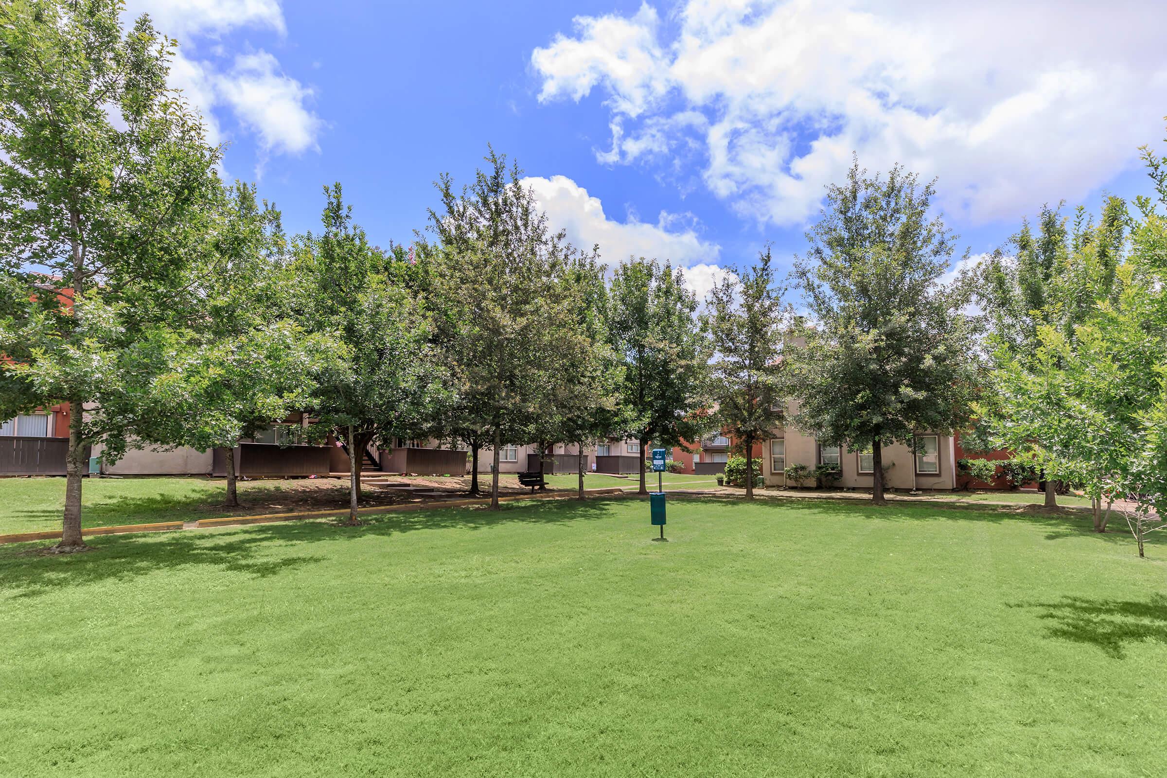 A spacious green lawn surrounded by well-maintained trees under a partly cloudy blue sky. In the center, there is a blue informational sign, and soft grass covers the ground, creating a serene outdoor environment.