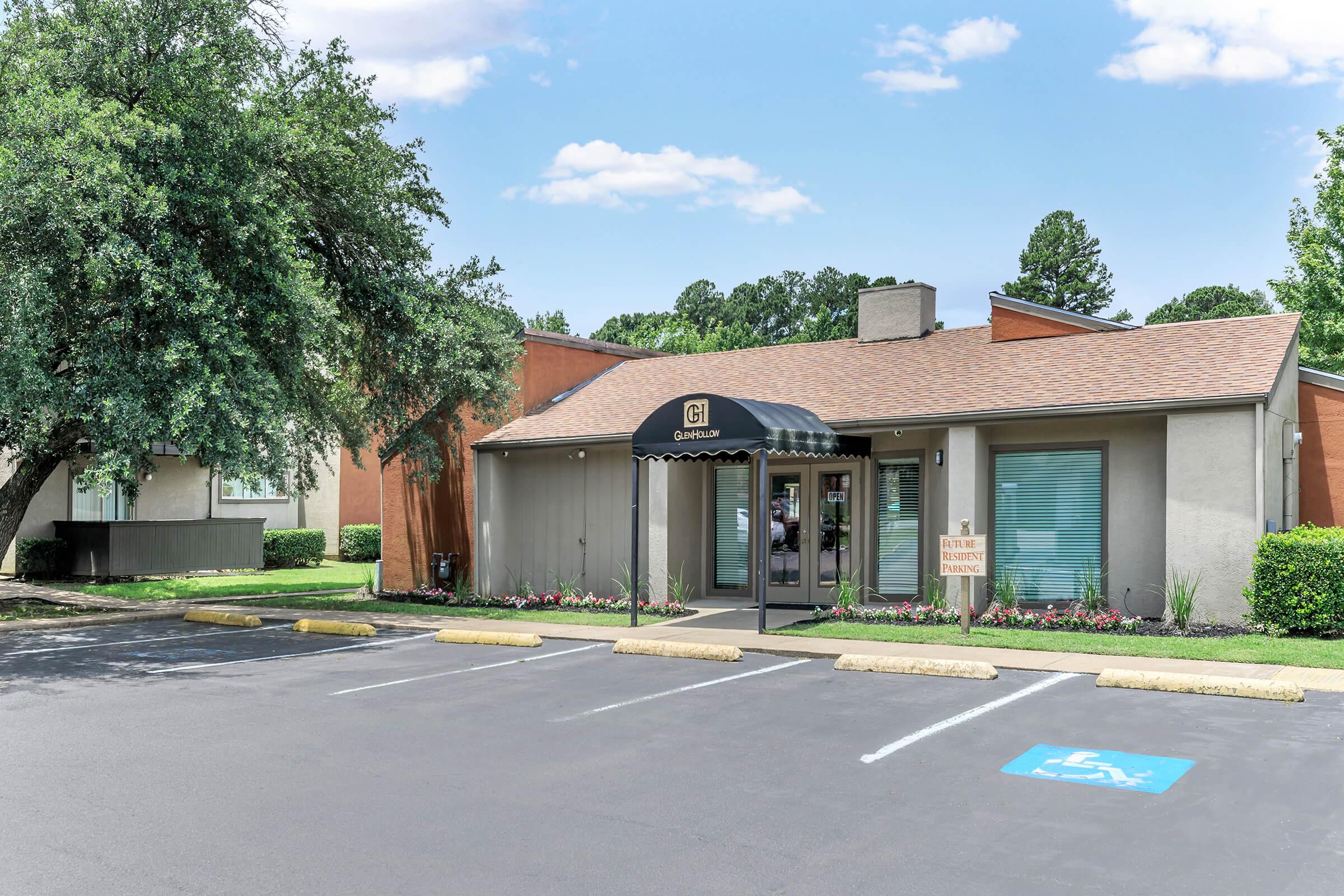 A modern building with a welcoming entrance under a black awning. The façade is light-colored, featuring large windows and landscaped flowerbeds. The surrounding area has green grass, trees, and a parking lot with marked spaces, including a designated spot for individuals with disabilities. Bright, clear sky overhead.