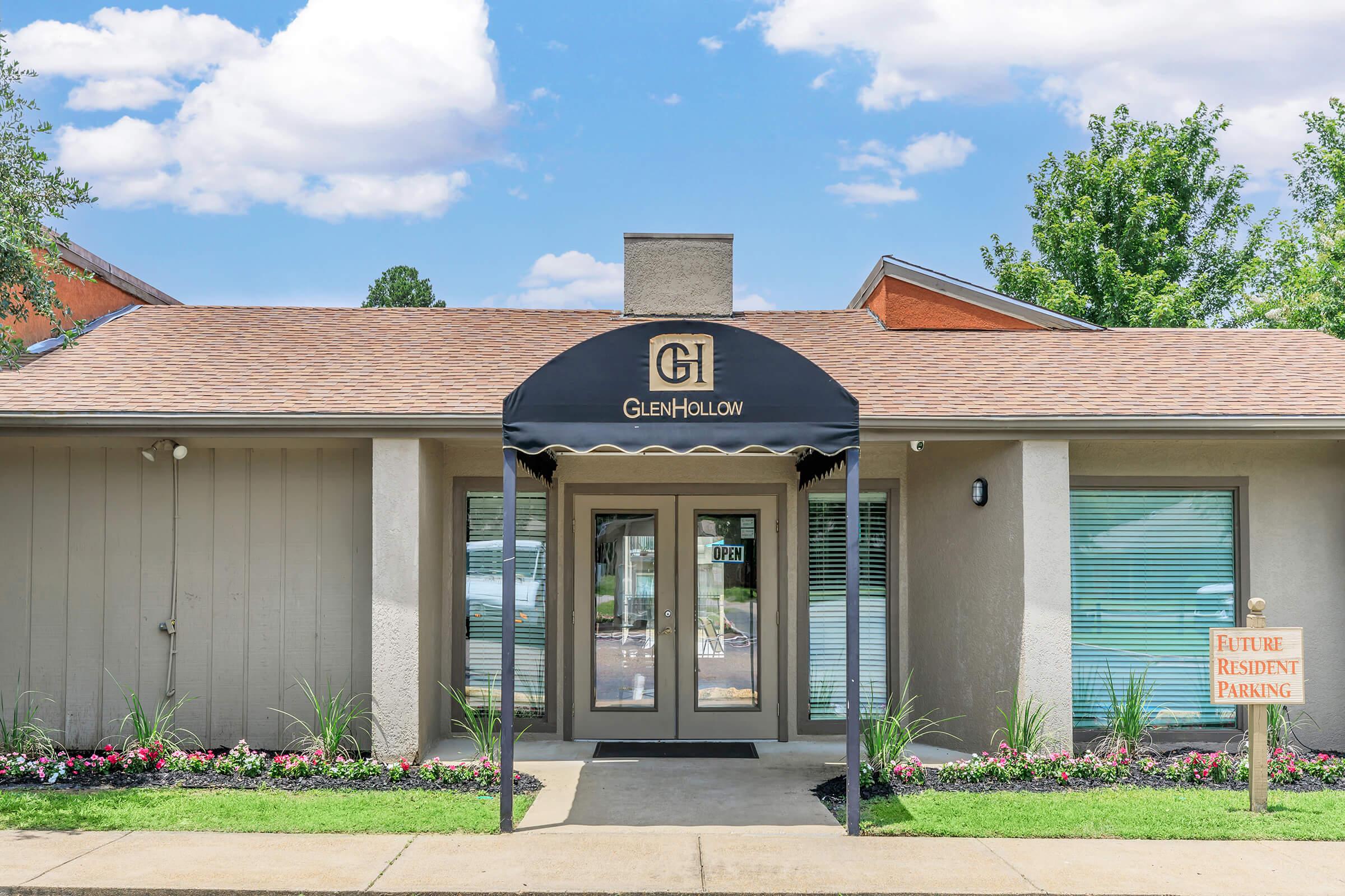 A building with a sign reading "Glen Hollow" above a covered entrance. The exterior features light-colored walls and a well-maintained landscape with flowers and grass. A "Future Resident Parking" sign is visible. The sky is partly cloudy, adding to the inviting atmosphere of the location.